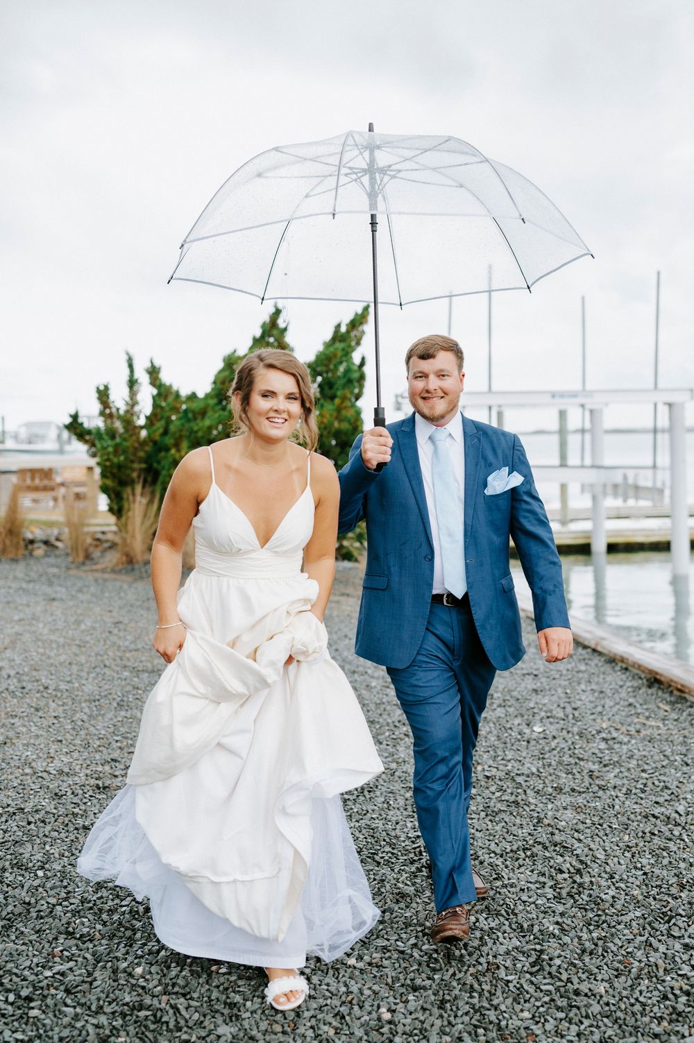 Smiling bride and groom walk together sharing a clear umbrella on a gravel path near a marina.
