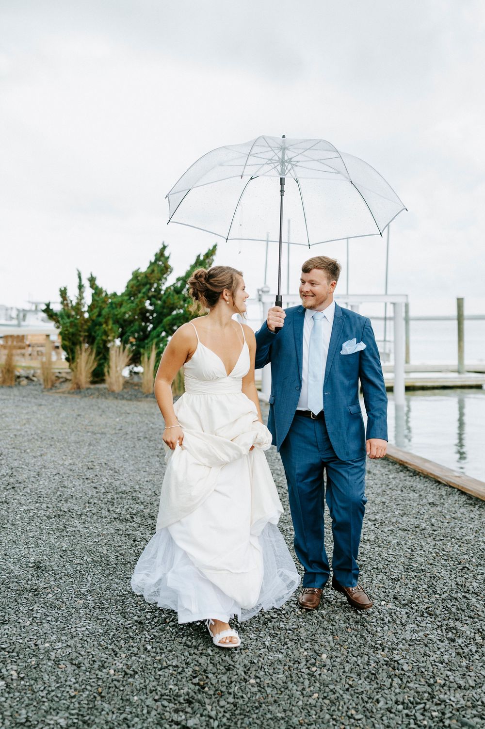 Smiling groom in navy suit holds clear umbrella over bride in white gown during outdoor waterfront wedding.