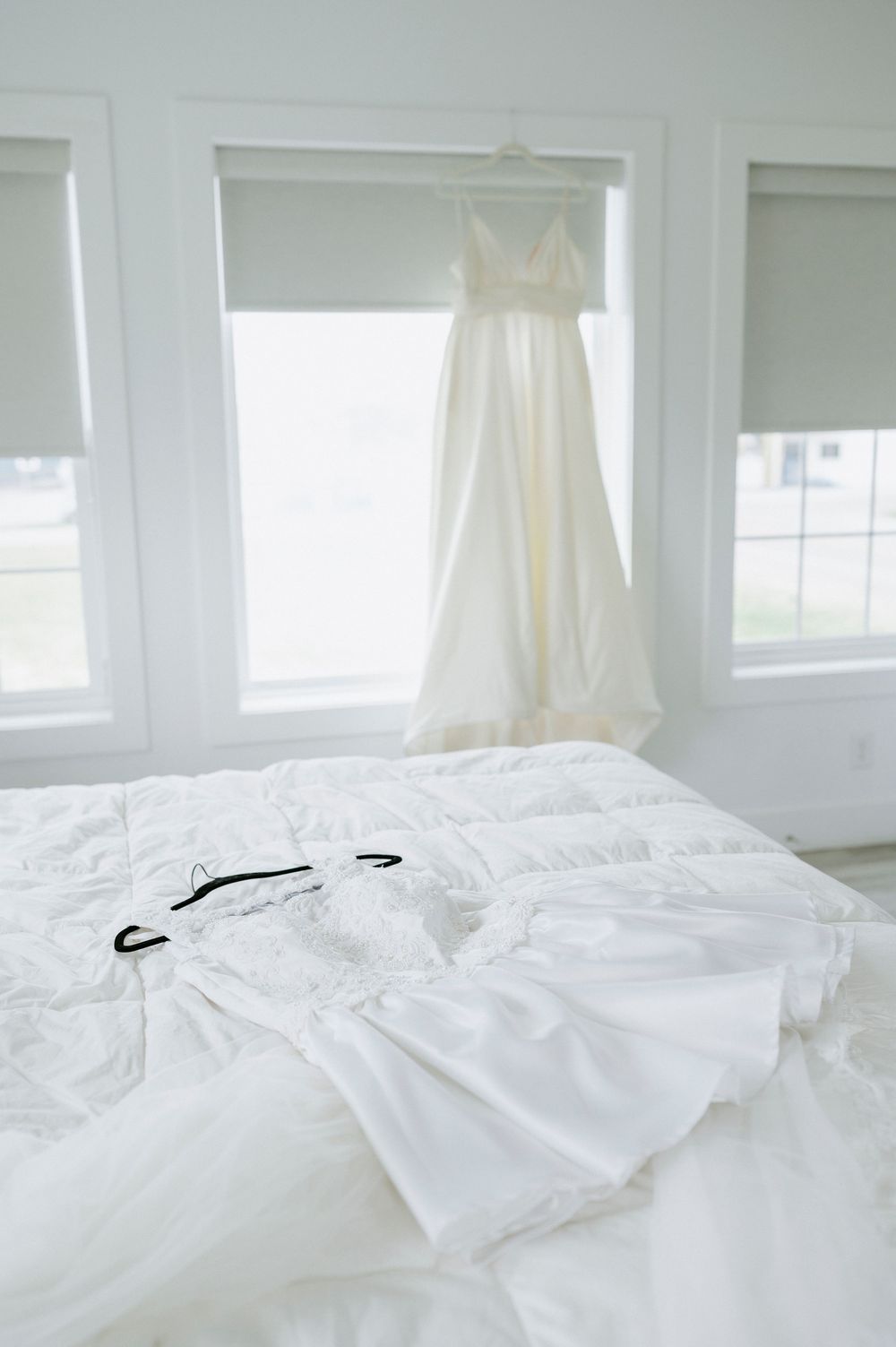 White wedding gown hangs in a bright window above a lace robe laid on a white bed.