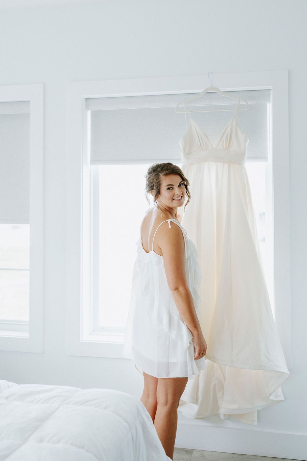 Bride in white slip dress smiles over shoulder at hanging wedding gown in bright white bedroom.