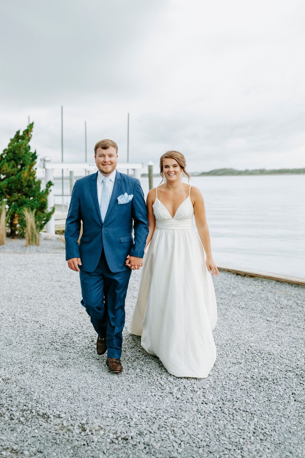 Happy bride and groom walk hand in hand along a gravel waterfront path, groom in blue suit, bride in elegant white dress.