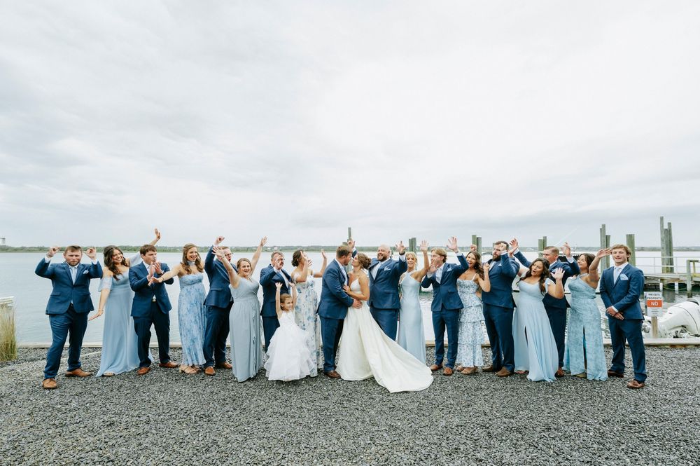 Large wedding party celebrating outdoors by the waterfront, bridesmaids in blue gowns and groomsmen in navy suits.
