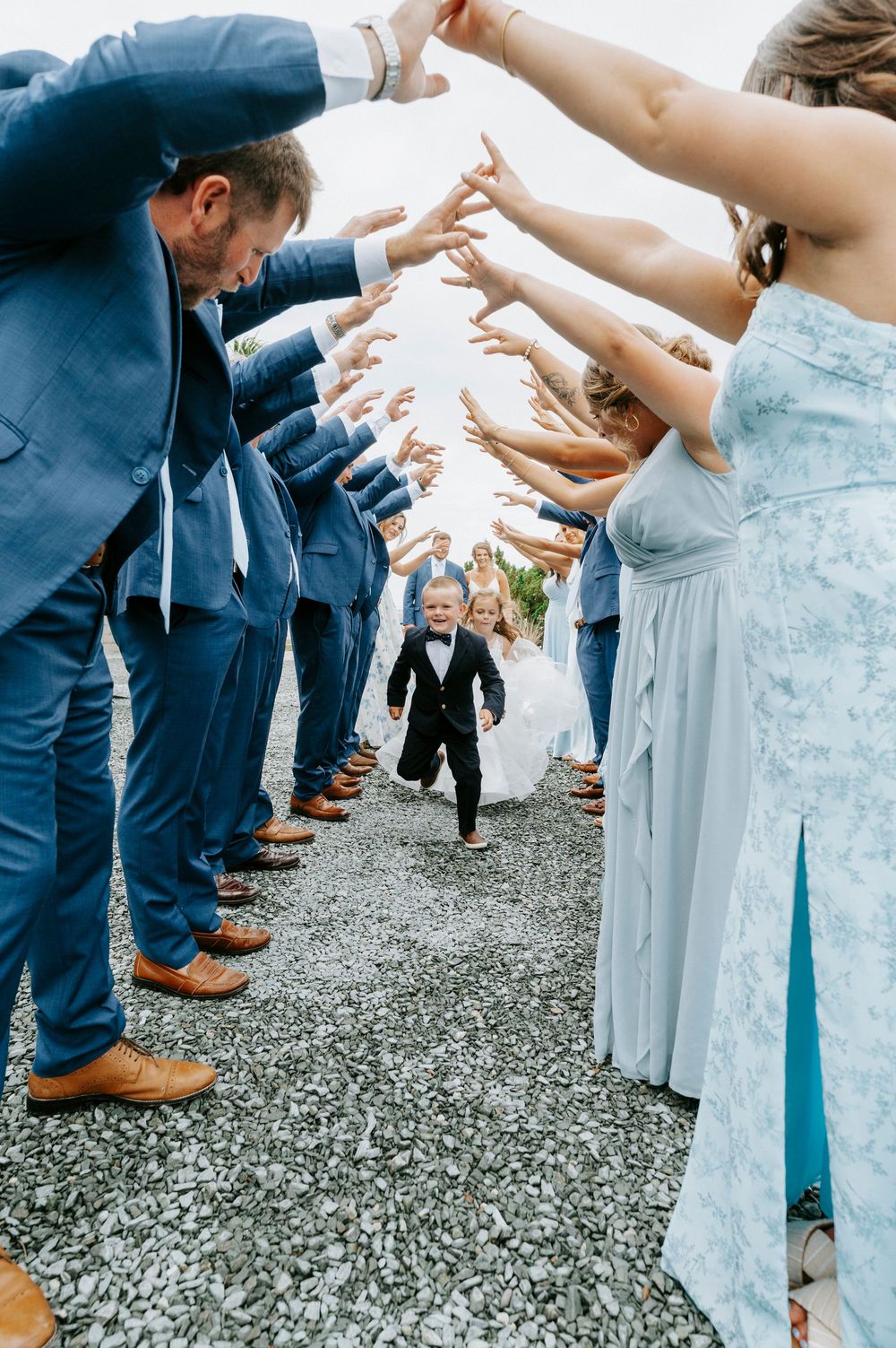 Excited toddler in a tuxedo runs through a tunnel of raised hands formed by the wedding party.