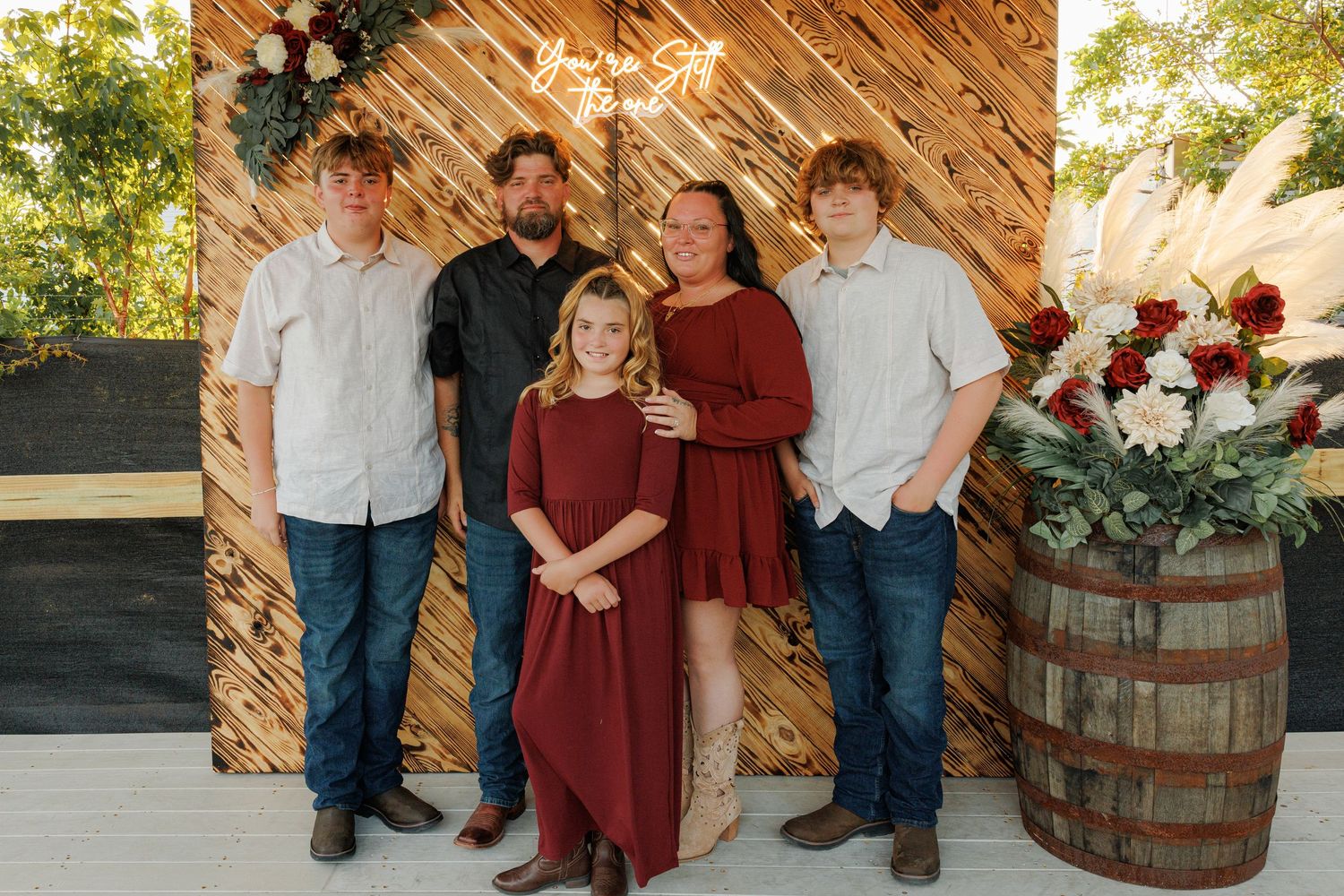 Family of five posing together in front of a rustic wooden backdrop with floral decorations and a barrel.