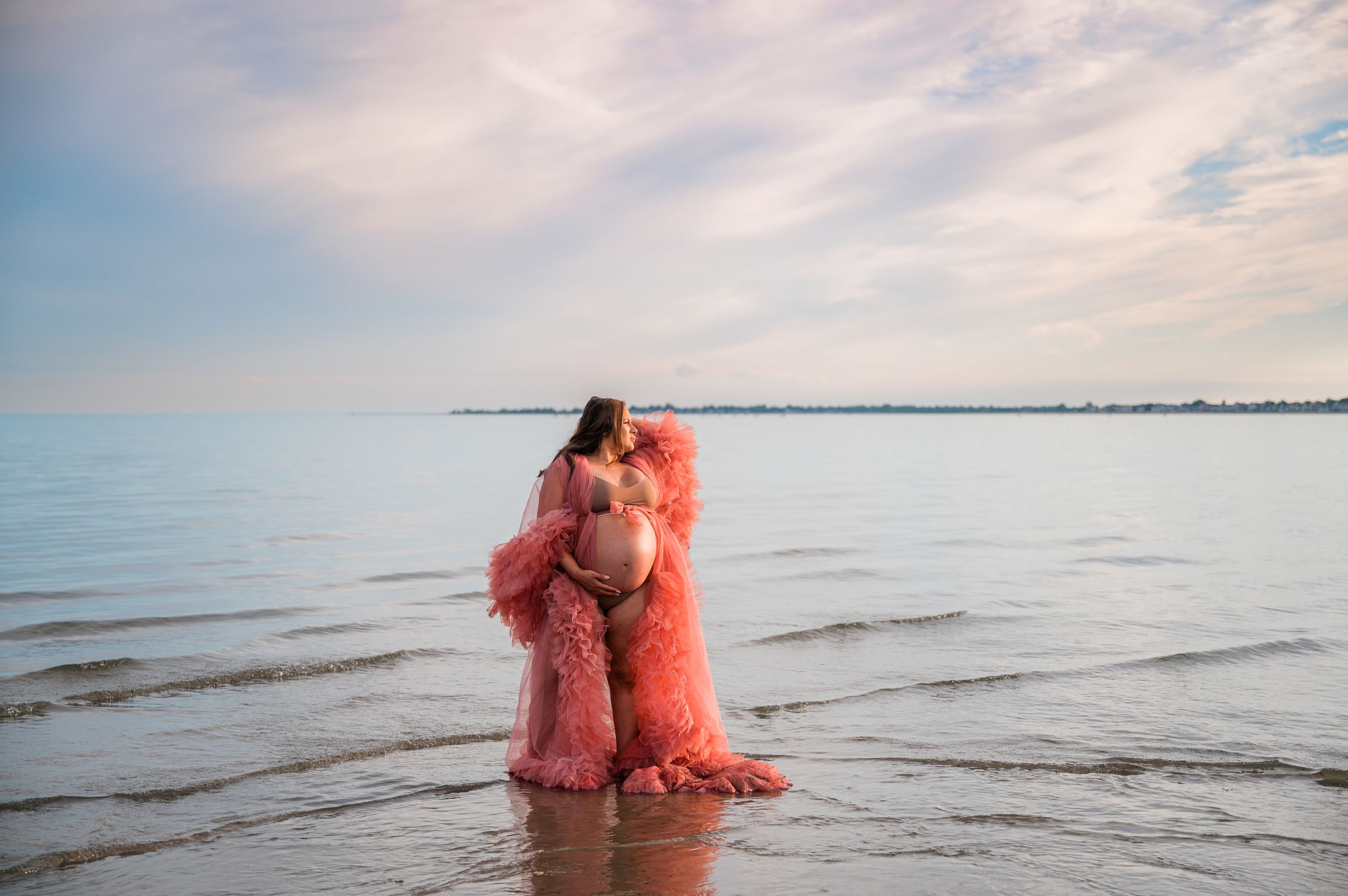 Pregnant woman in pink feather robe standing in calm ocean water at sunset with cloudy sky backdrop in Milford CT.