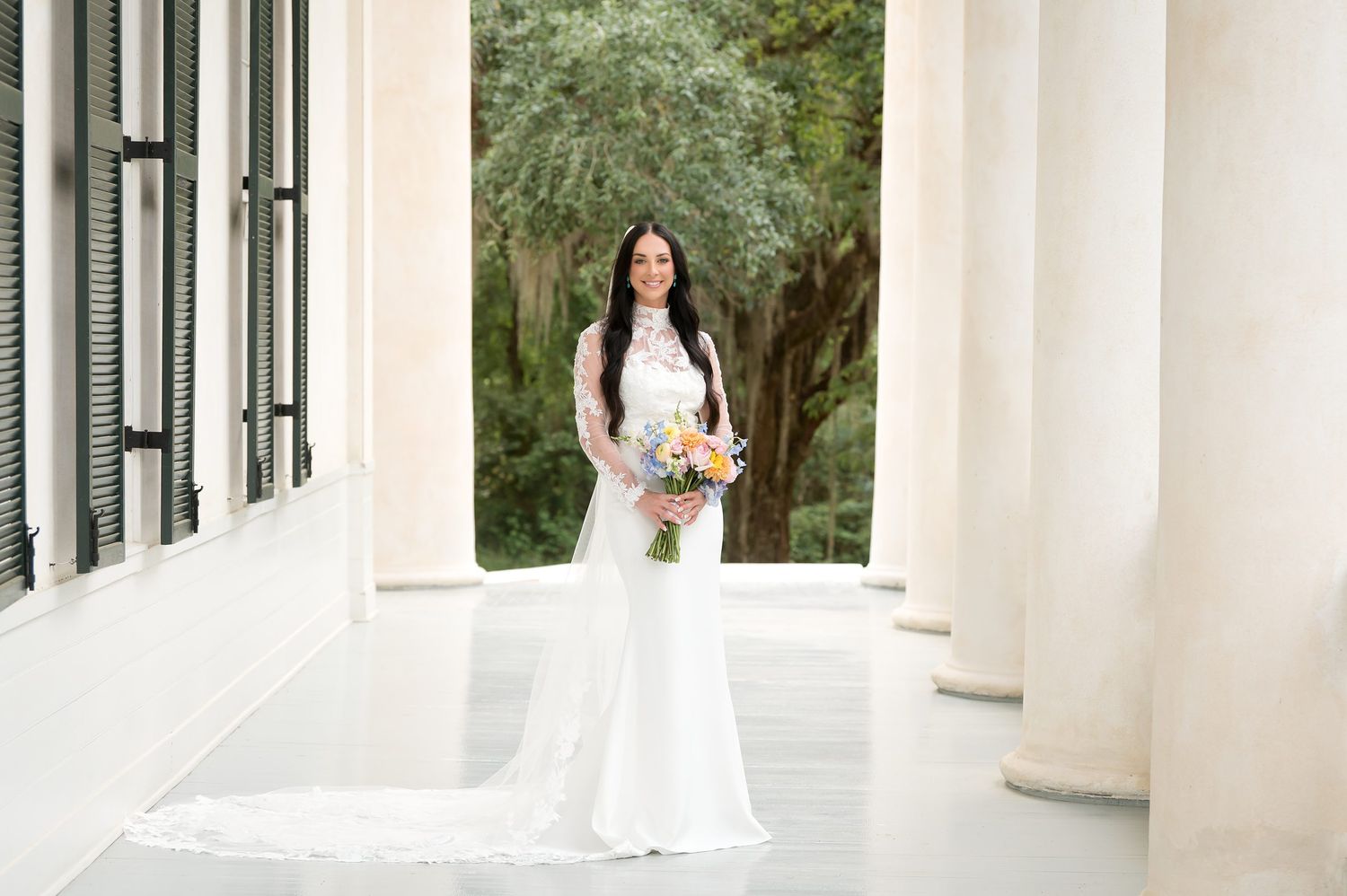 Bride in white lace gown holding colorful bouquet standing between elegant columns at outdoor venue.