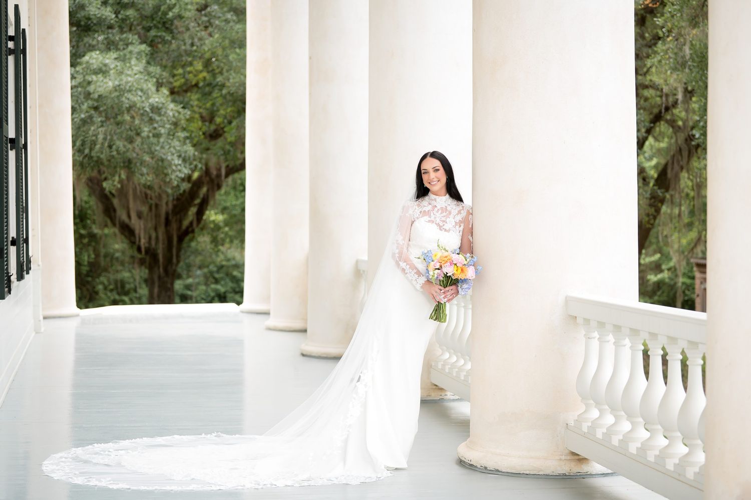 Bride in elegant white gown with long train holds bouquet on classical columned porch surrounded by lush greenery.