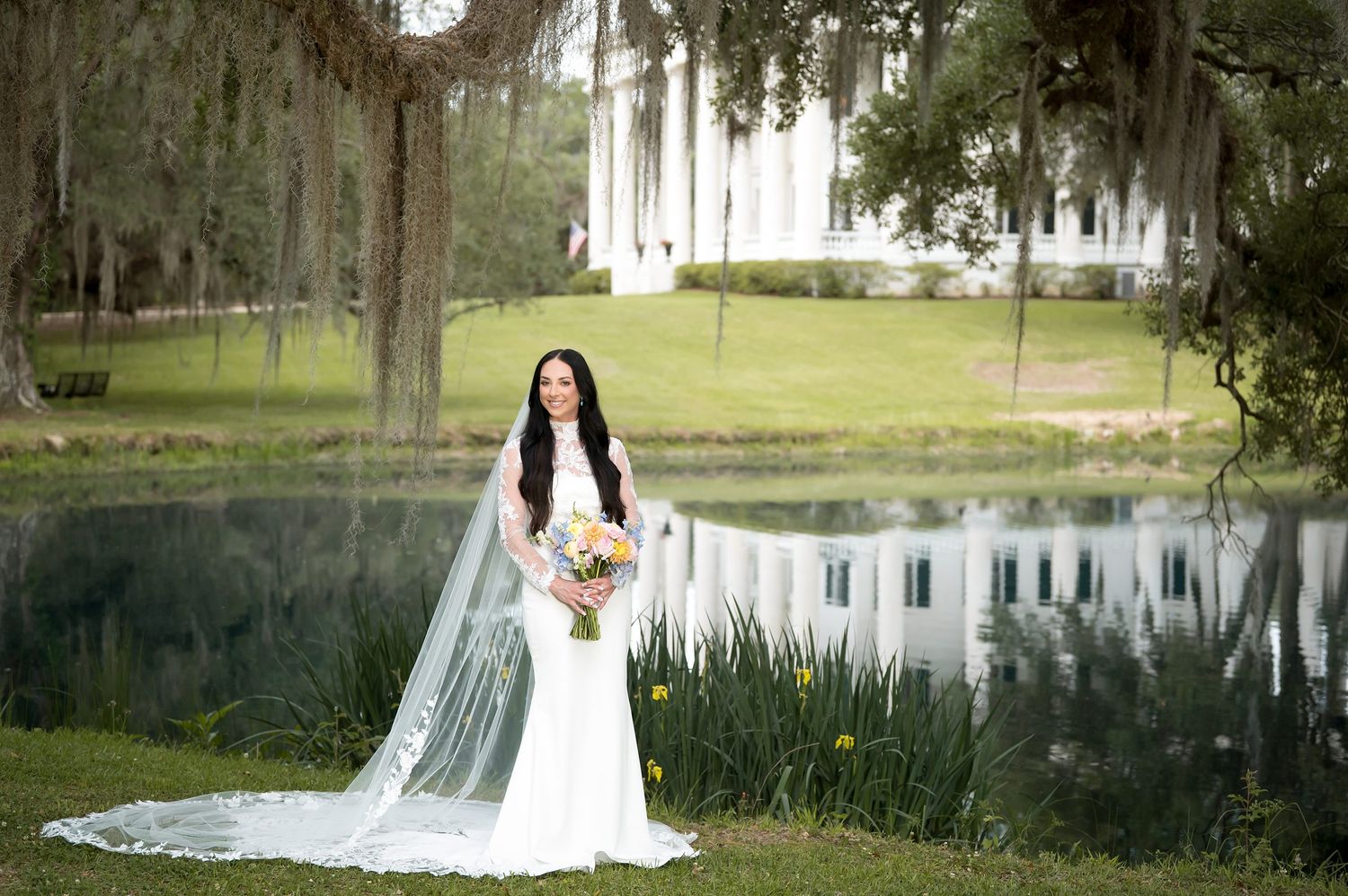 Bride in white gown with long veil holds bouquet by pond at Southern plantation wedding venue.
