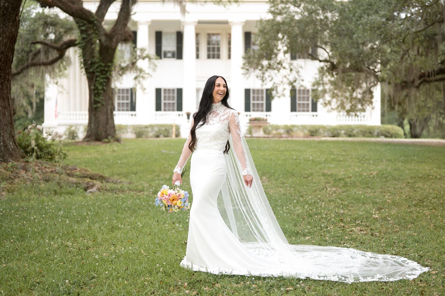 Bride in elegant white gown with sheer cape holds colorful bouquet on green lawn before white plantation mansion.