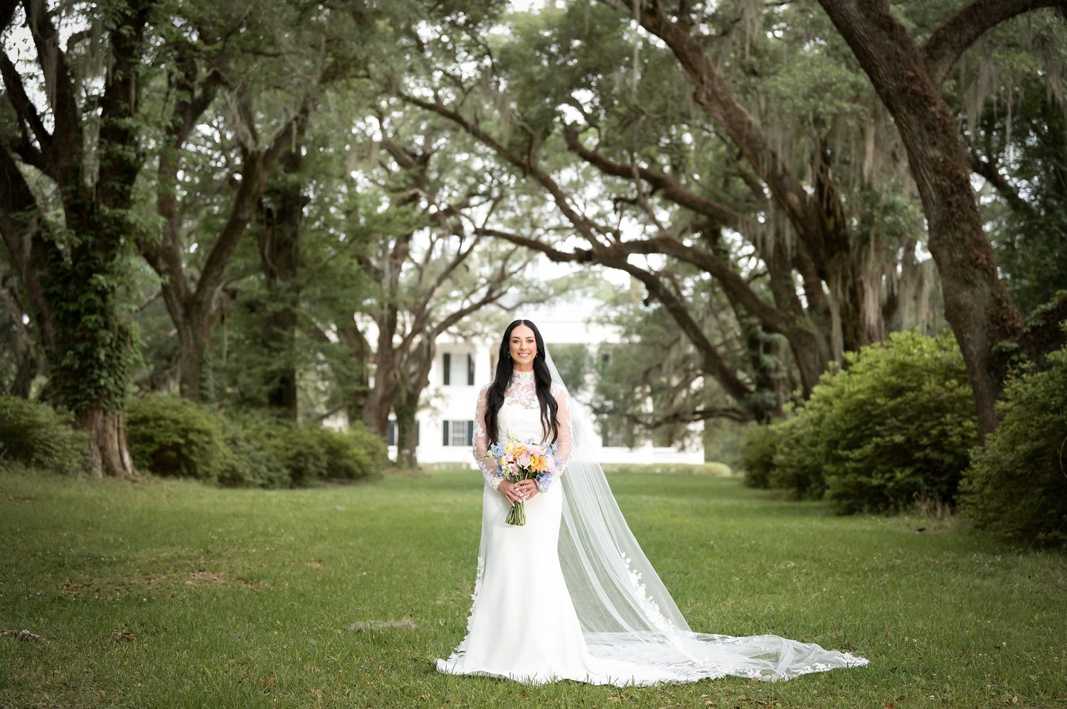 Bride in white gown with long veil holds bouquet on green lawn with moss-draped oak trees and white plantation house.