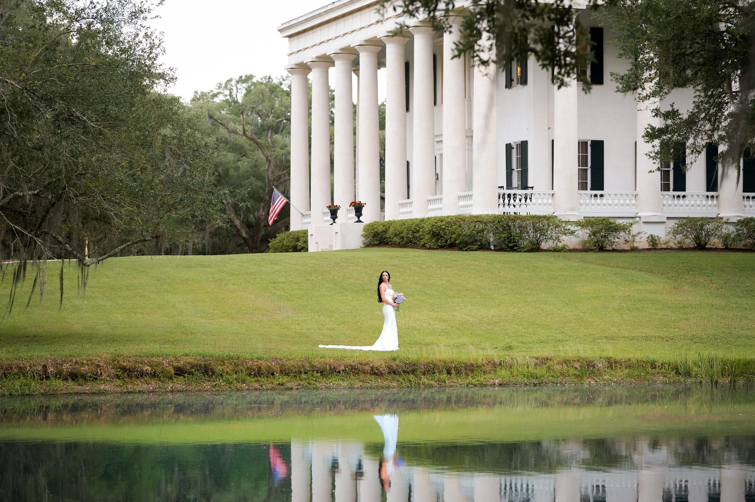 Bride and groom embrace on lush green lawn before grand white Southern plantation mansion with reflecting pond.