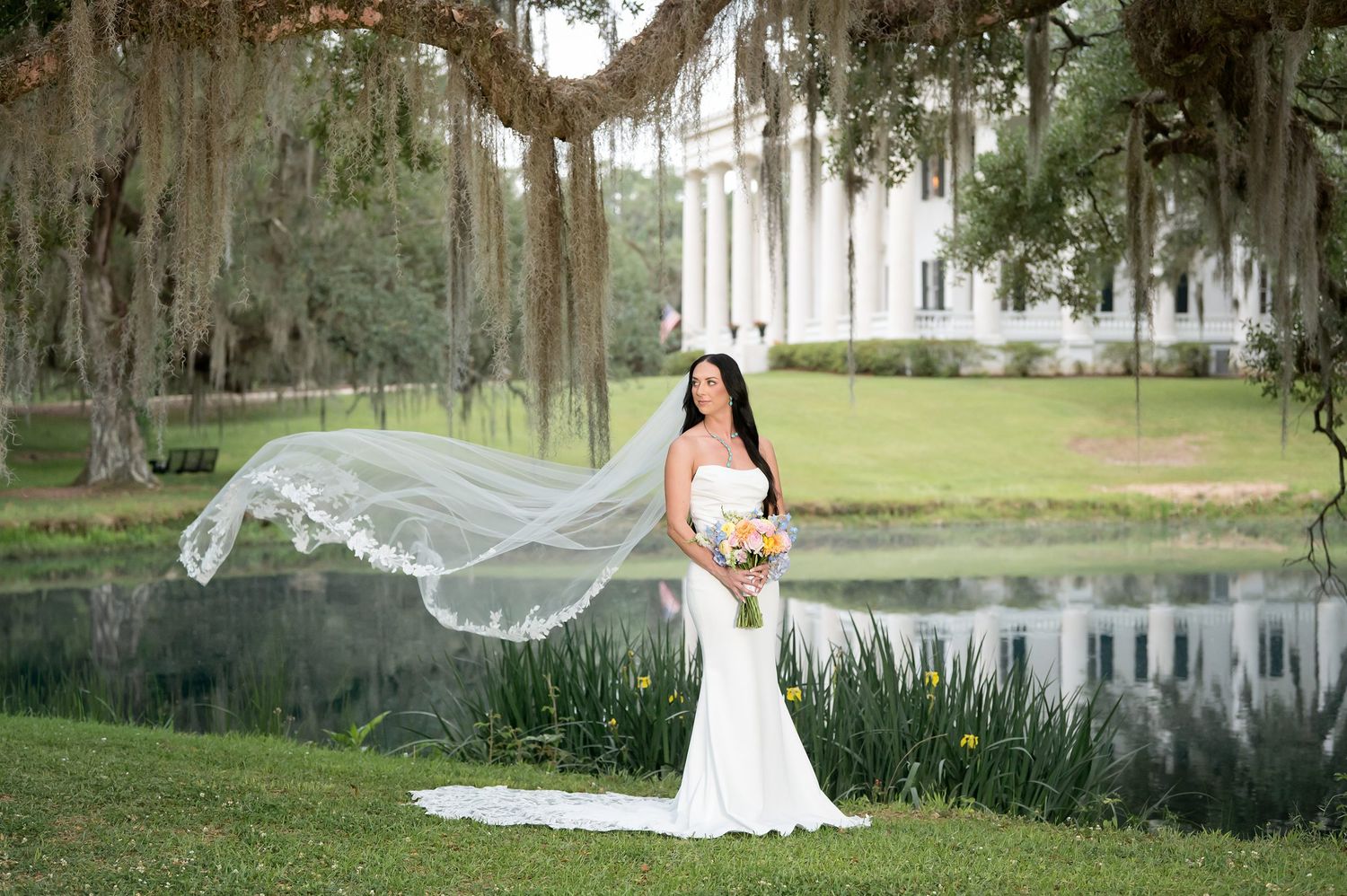 Bride in white mermaid gown with flowing veil by pond under Spanish moss trees at Southern plantation.
