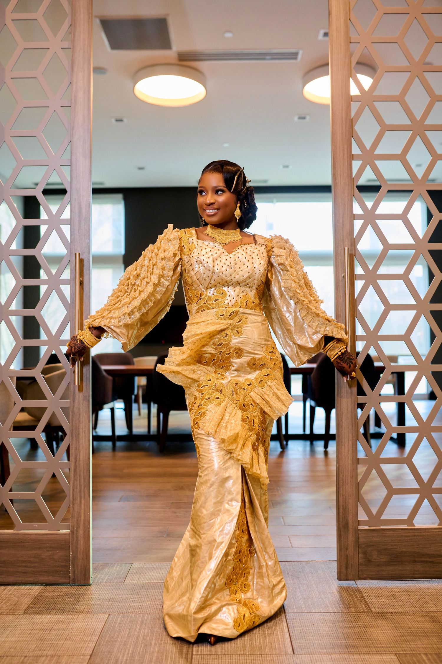 Woman in elegant gold African traditional dress posing gracefully in a modern interior doorway.