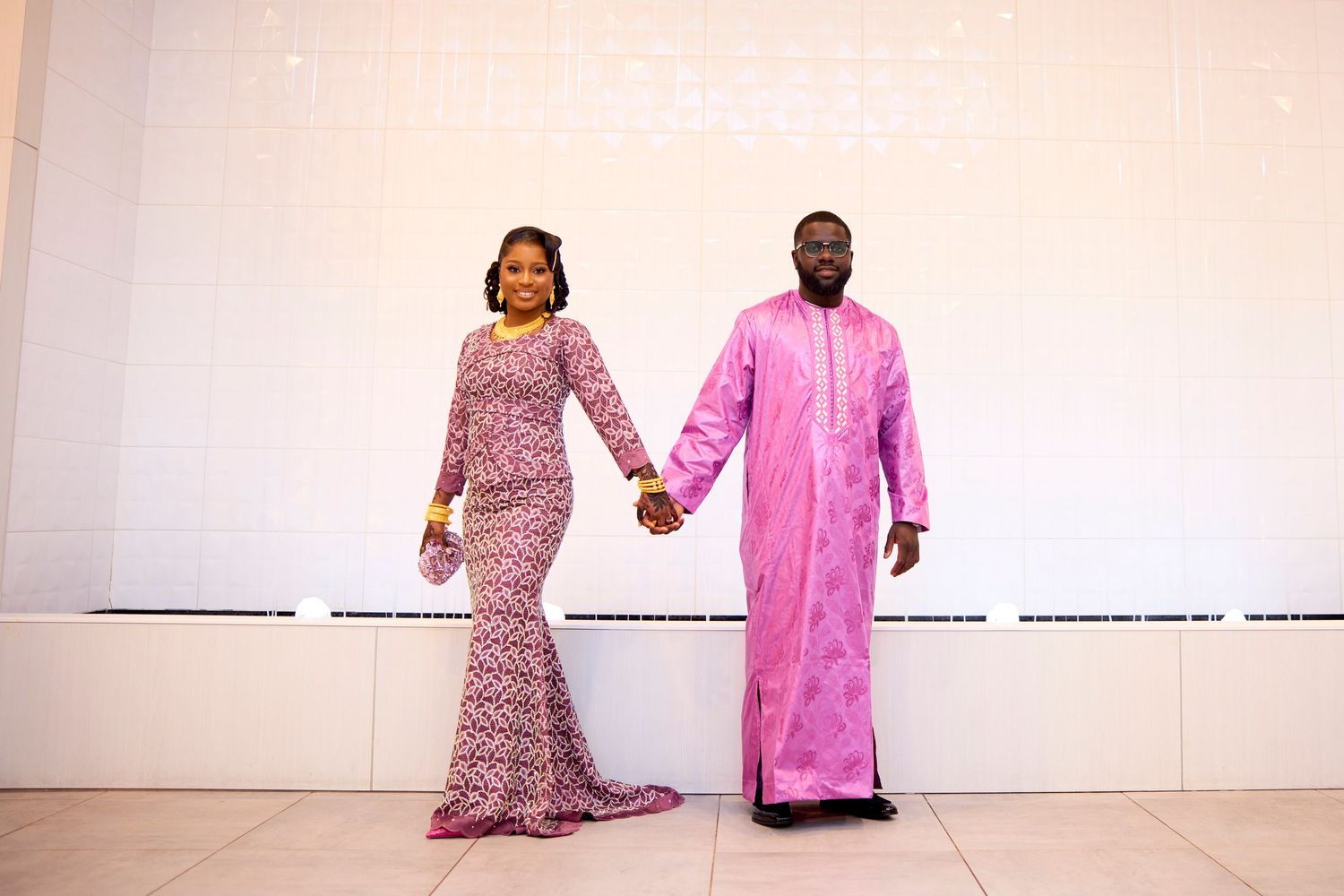Couple in matching pink outfits holding hands, wearing elegant African attire, posing together against a white wall.