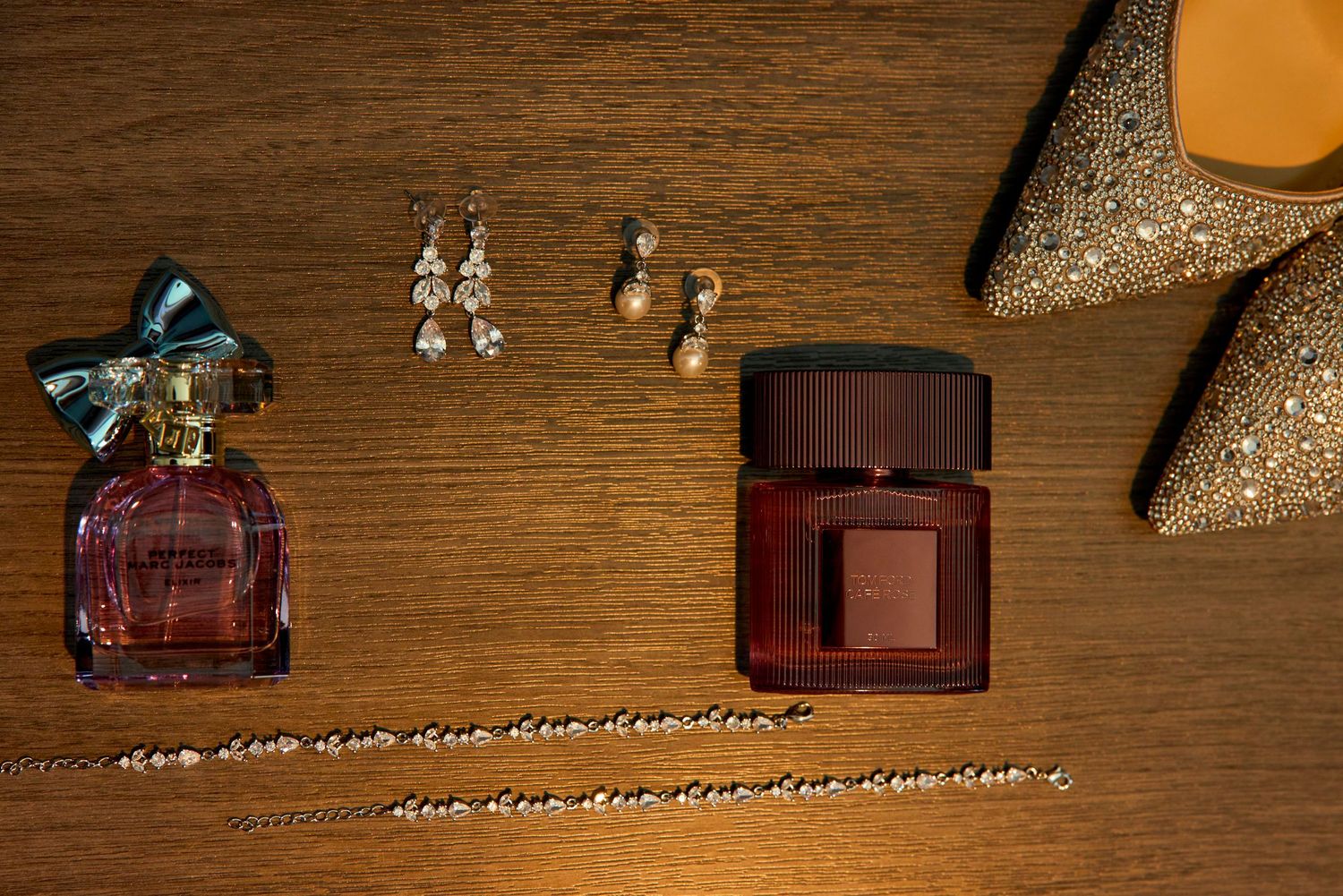 Elegant flat lay of two perfume bottles, pearl drop earrings, a gold necklace, and glittery heels on wooden surface.