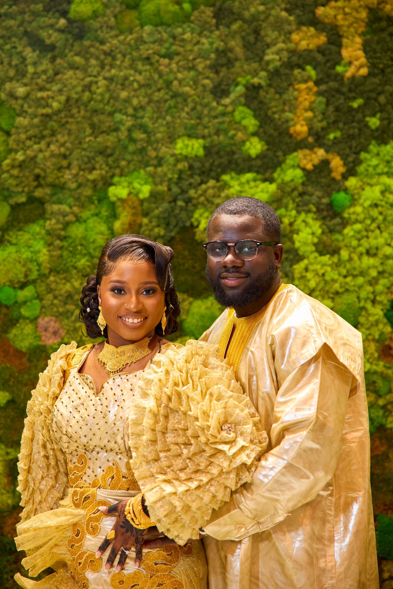 Couple in gold traditional African attire posing together in front of a lush green moss wall backdrop.
