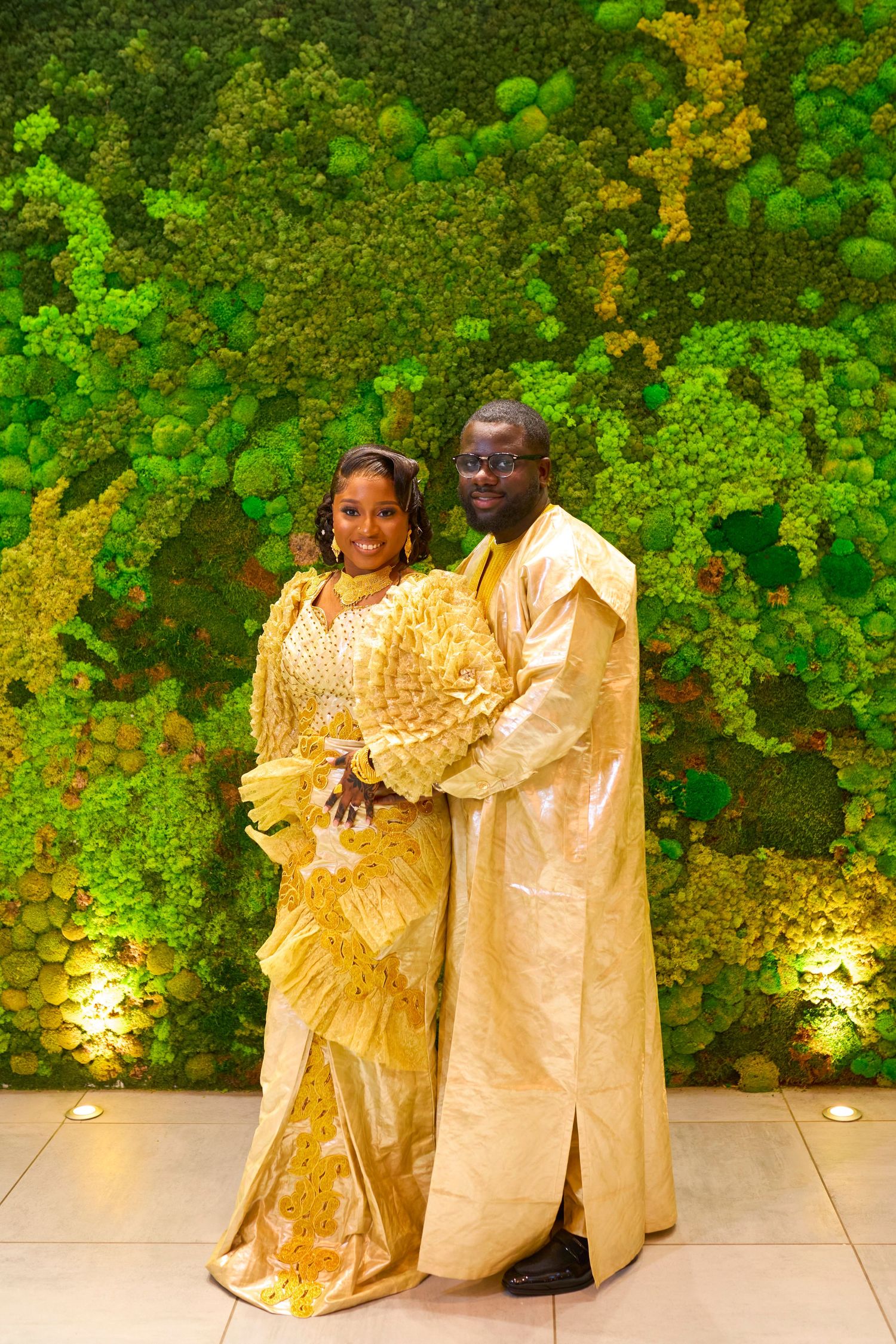 Couple in elegant gold African traditional attire posing before a lush green moss wall backdrop.