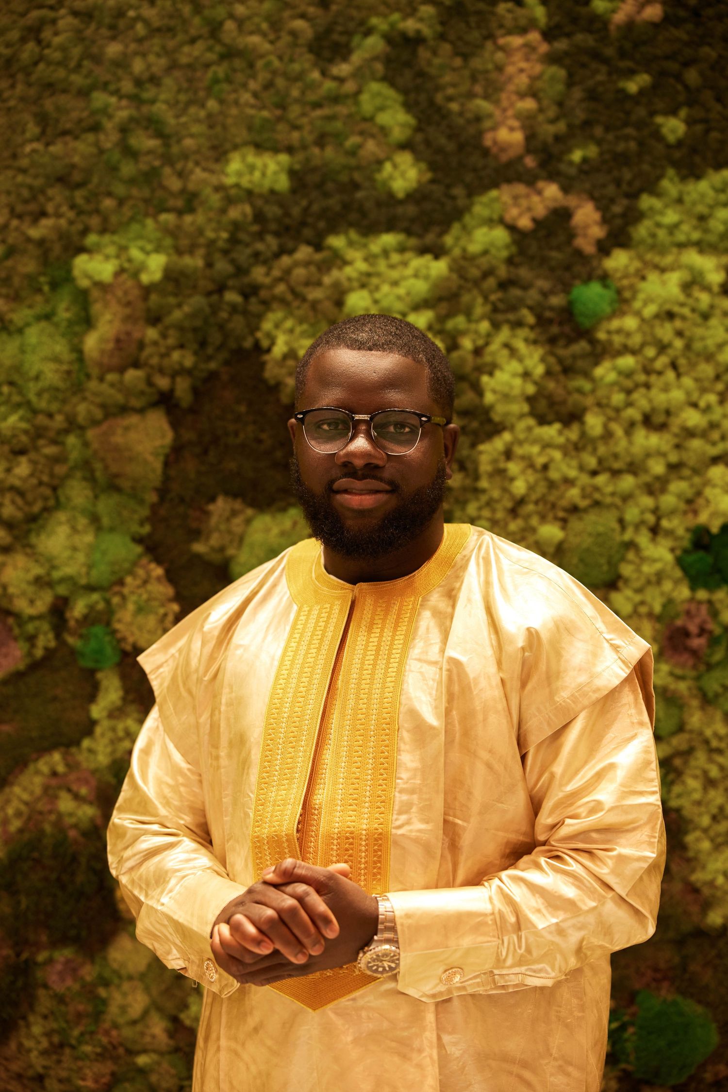 Man in gold traditional African attire and glasses poses confidently before a lush green moss wall backdrop.