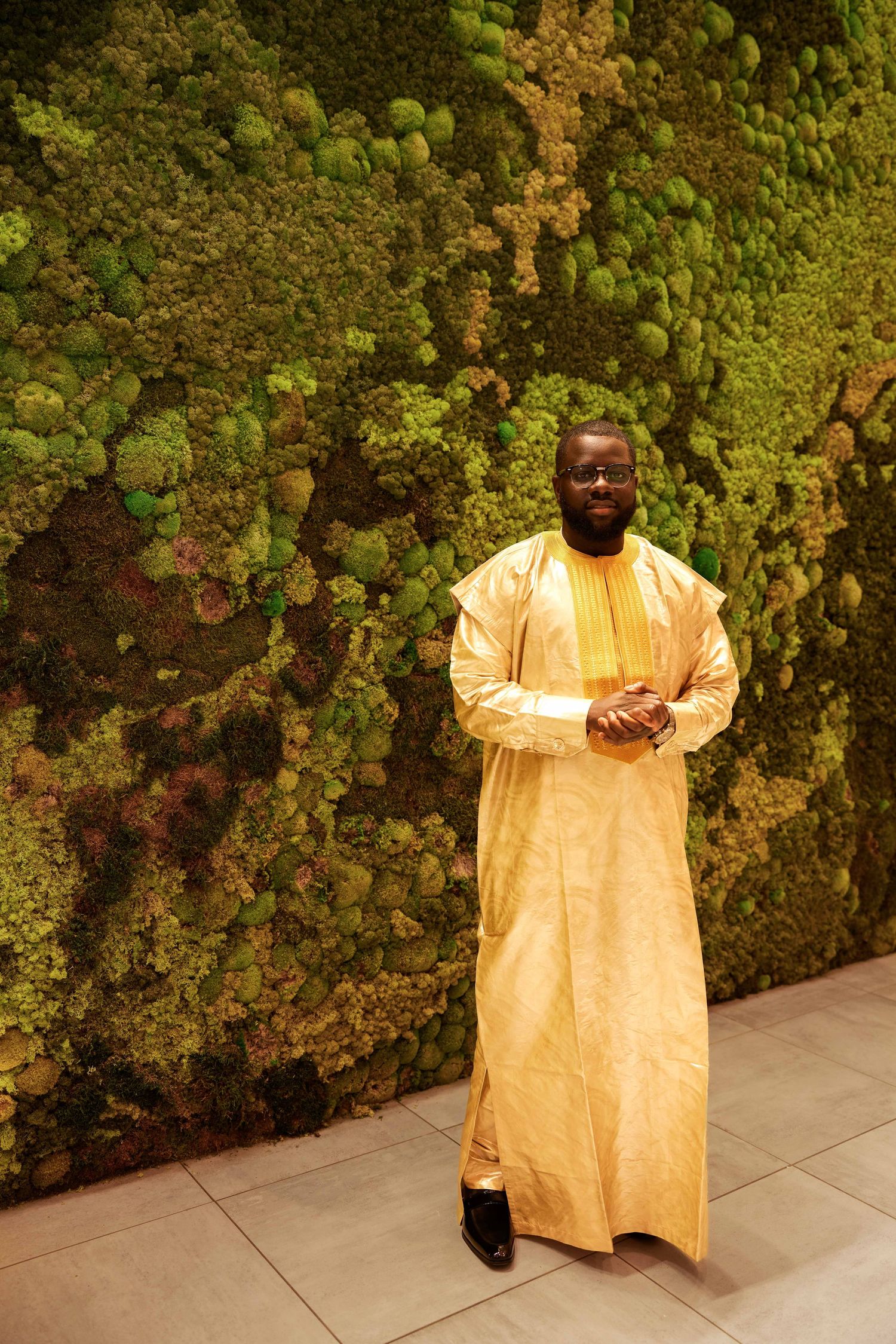 Man in gold African traditional attire standing before lush green moss wall.
