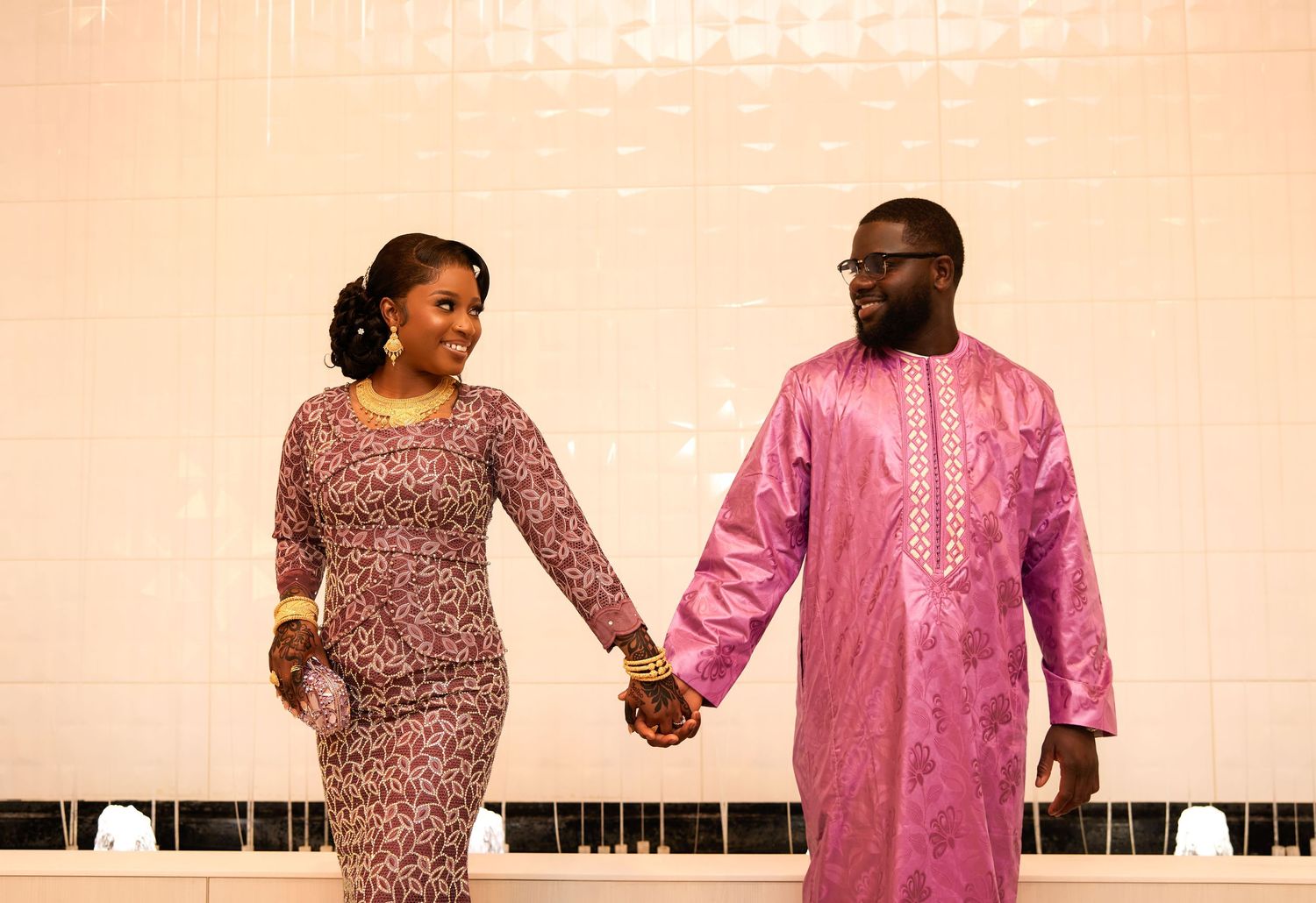 Couple in matching pink traditional African attire holding hands, smiling at each other in elegant indoor setting.