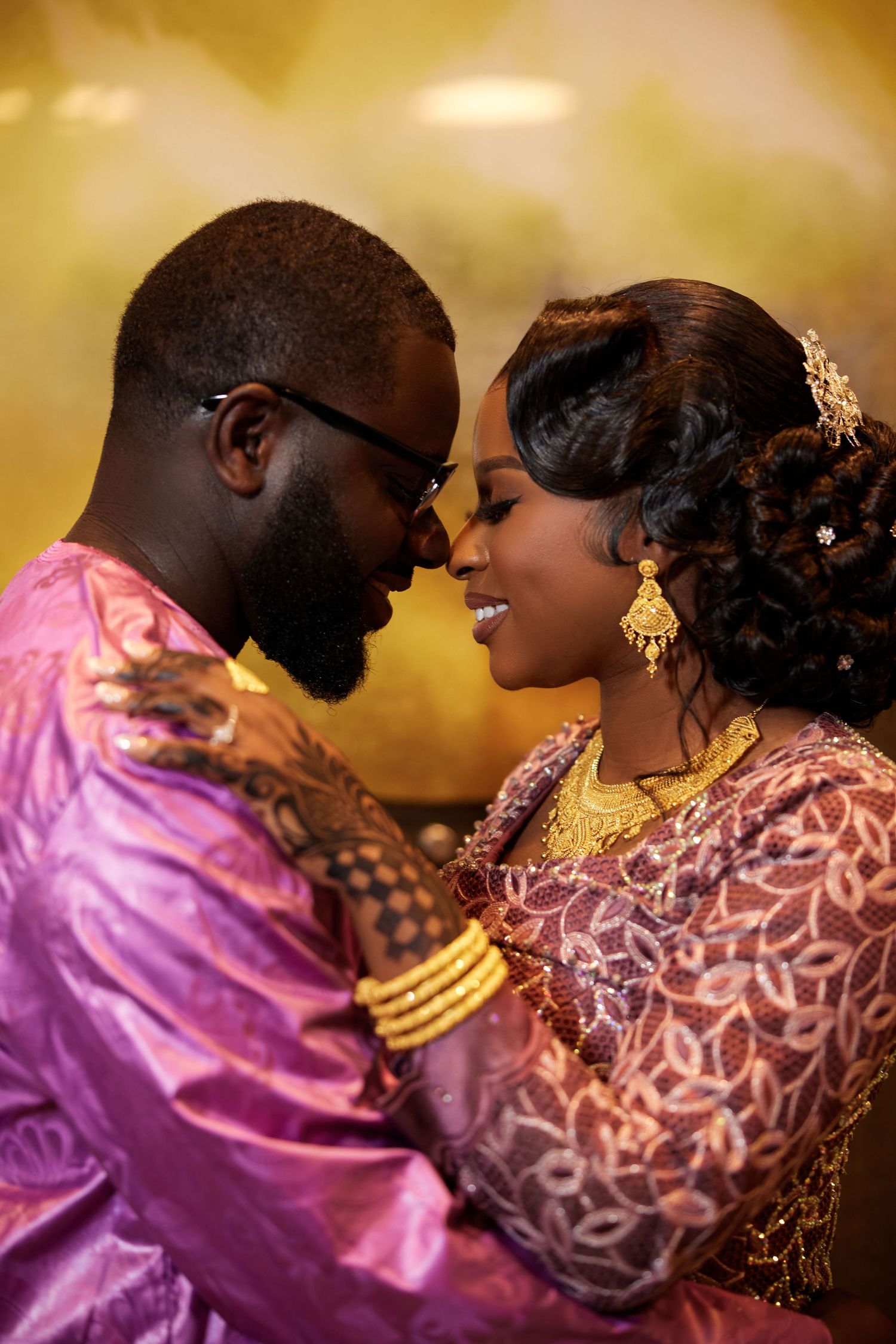 A couple in traditional African wedding attire share an intimate moment, adorned with gold jewelry and intricate henna designs.