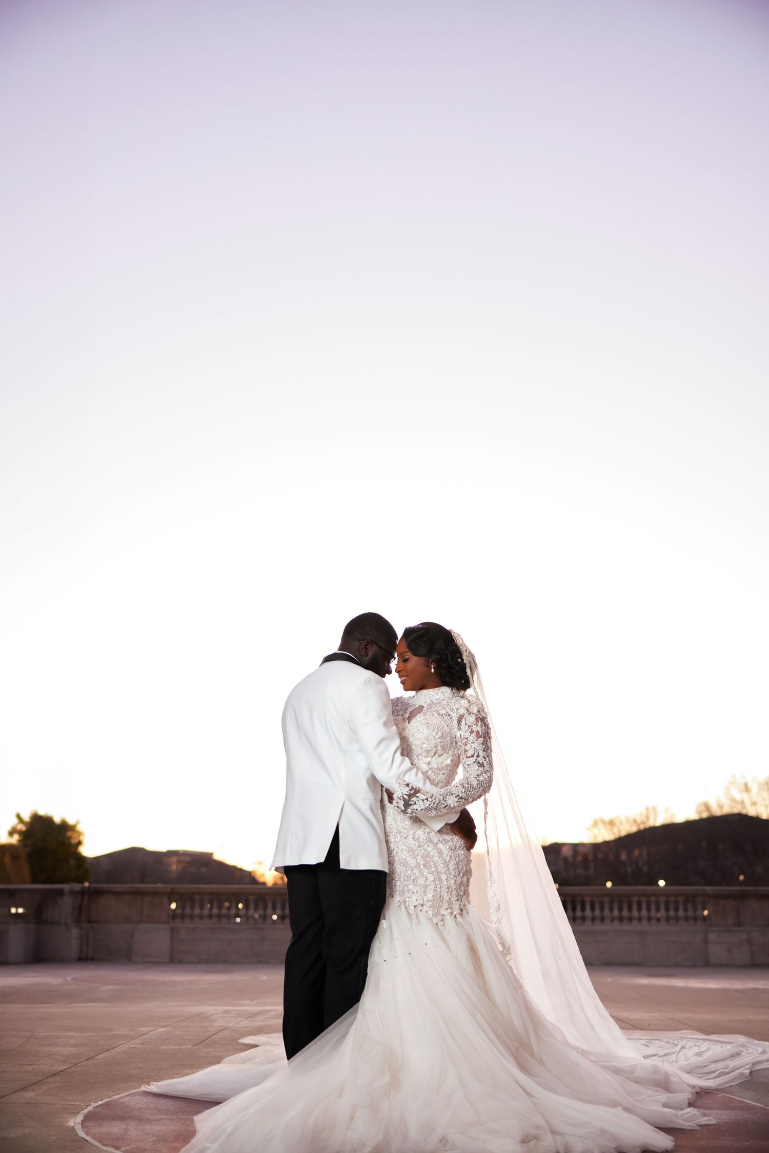 A bride and groom share a romantic embrace at sunset, the bride wearing a lace mermaid gown with a long veil.