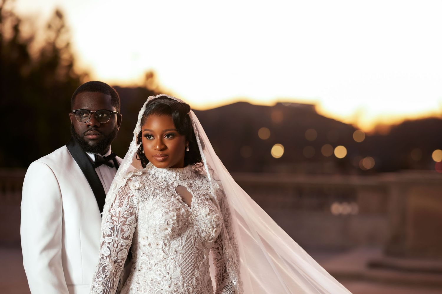 Elegant bride and groom posing at sunset, bride wearing lace long-sleeve gown with veil, groom in white tuxedo jacket.