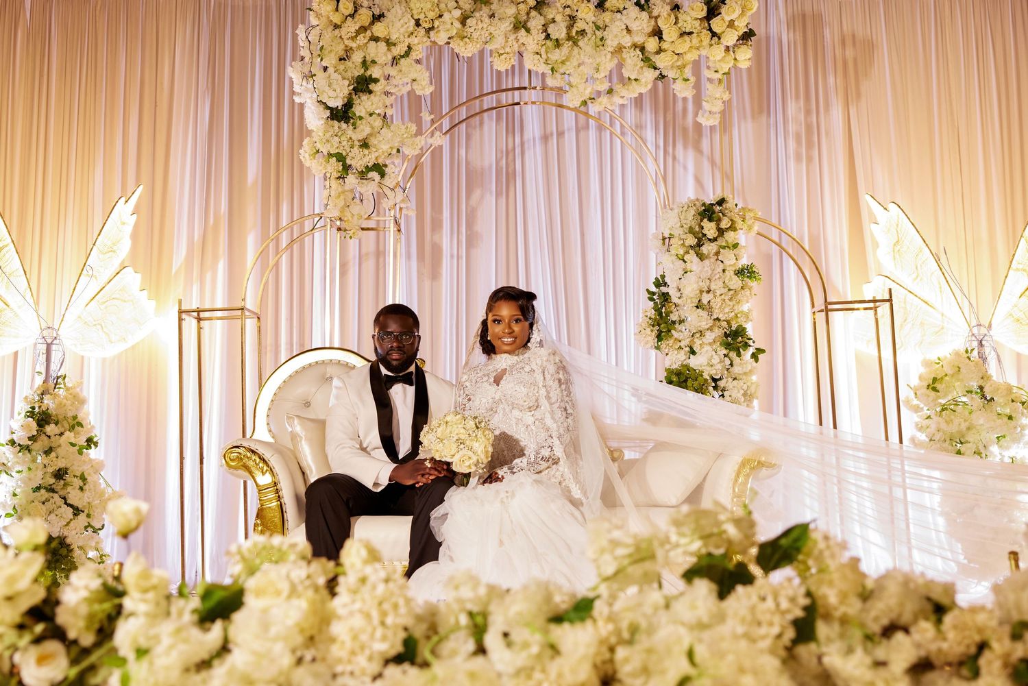 Elegant wedding couple seated on gold sofa amid white floral arch and draping backdrop at luxury reception.