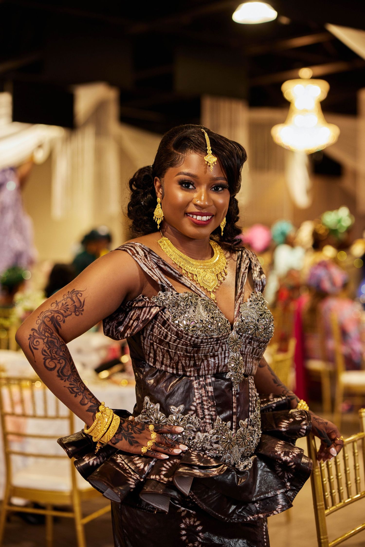 Woman in ornate African attire with gold jewelry and henna tattoo smiles at a festive cultural celebration event.