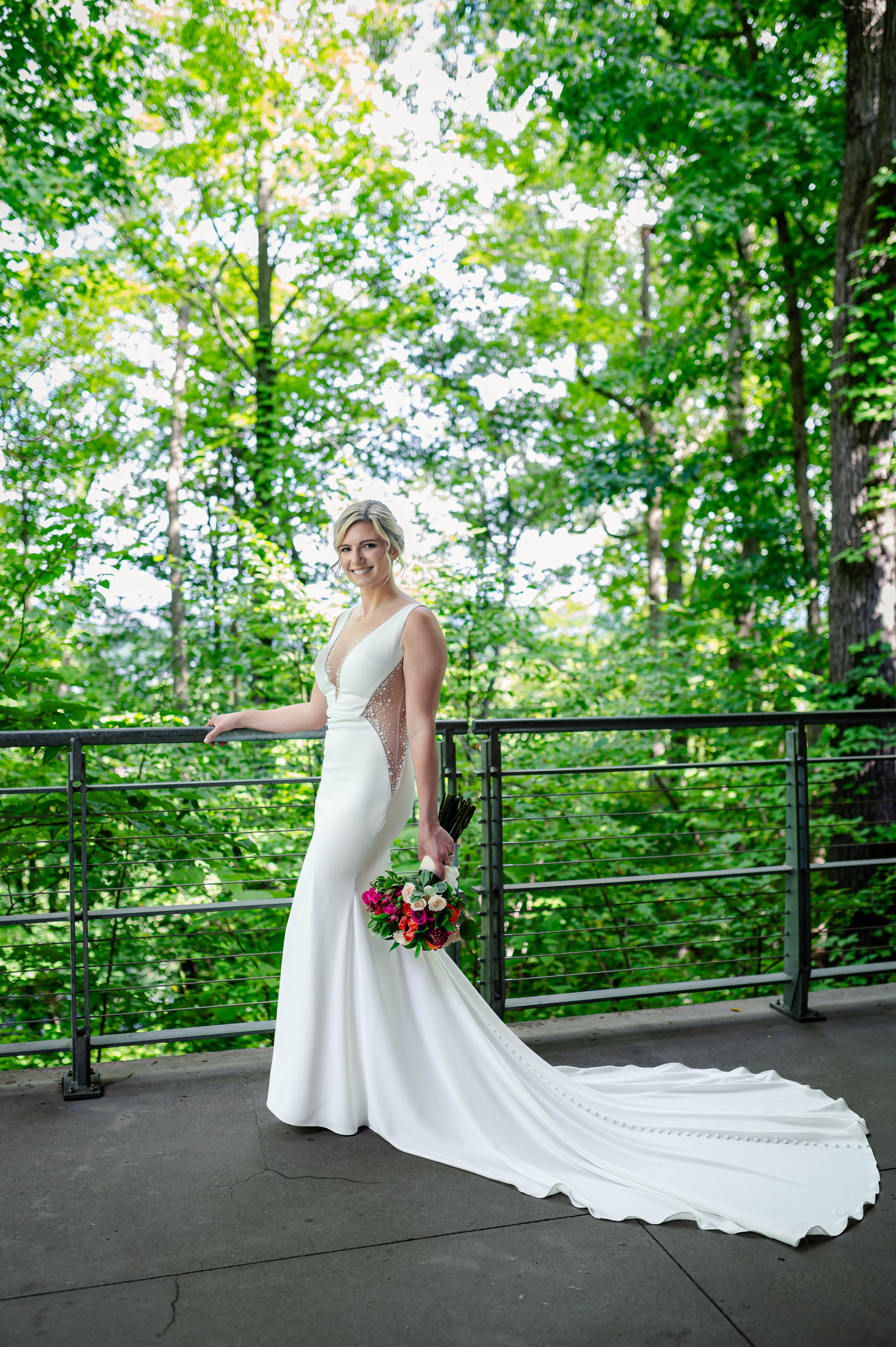 Bride in elegant white mermaid gown with train poses on outdoor balcony surrounded by lush green trees.