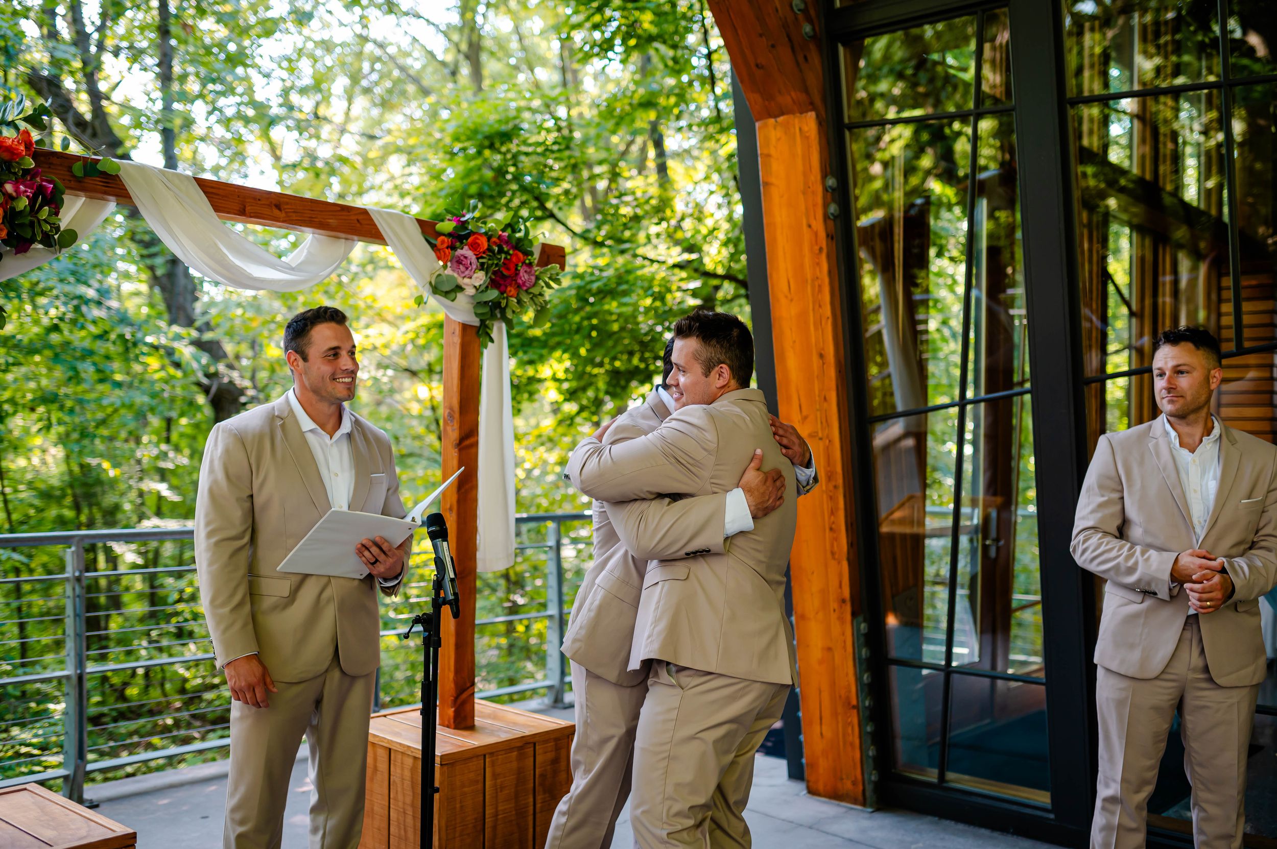 Groomsmen in tan suits share a hug during an outdoor wedding ceremony under a floral arch.