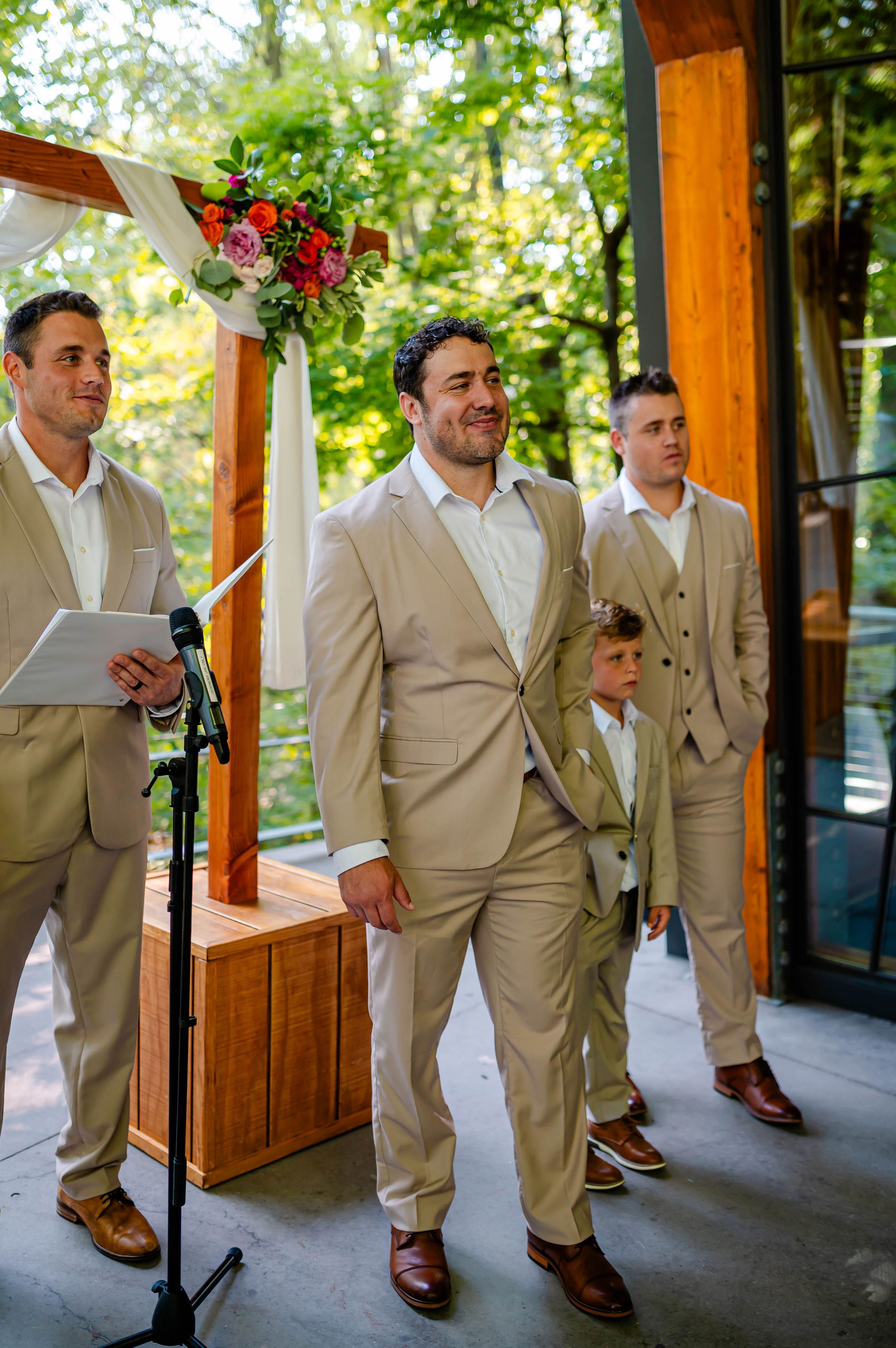 Groom in tan suit smiles at altar with groomsmen and ring bearer during outdoor wedding ceremony.