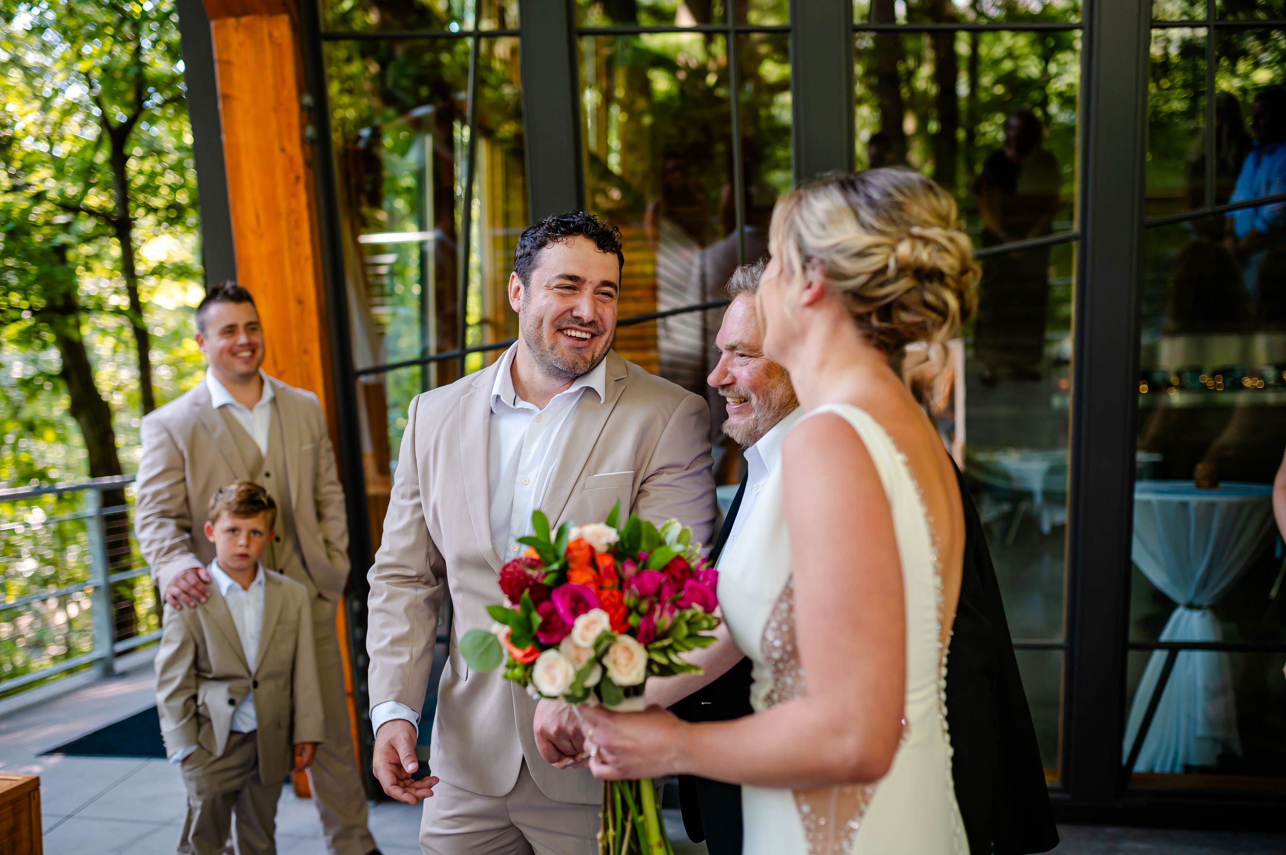 Groom in tan suit smiling at bride holding colorful bouquet at outdoor wedding venue with guests in background.