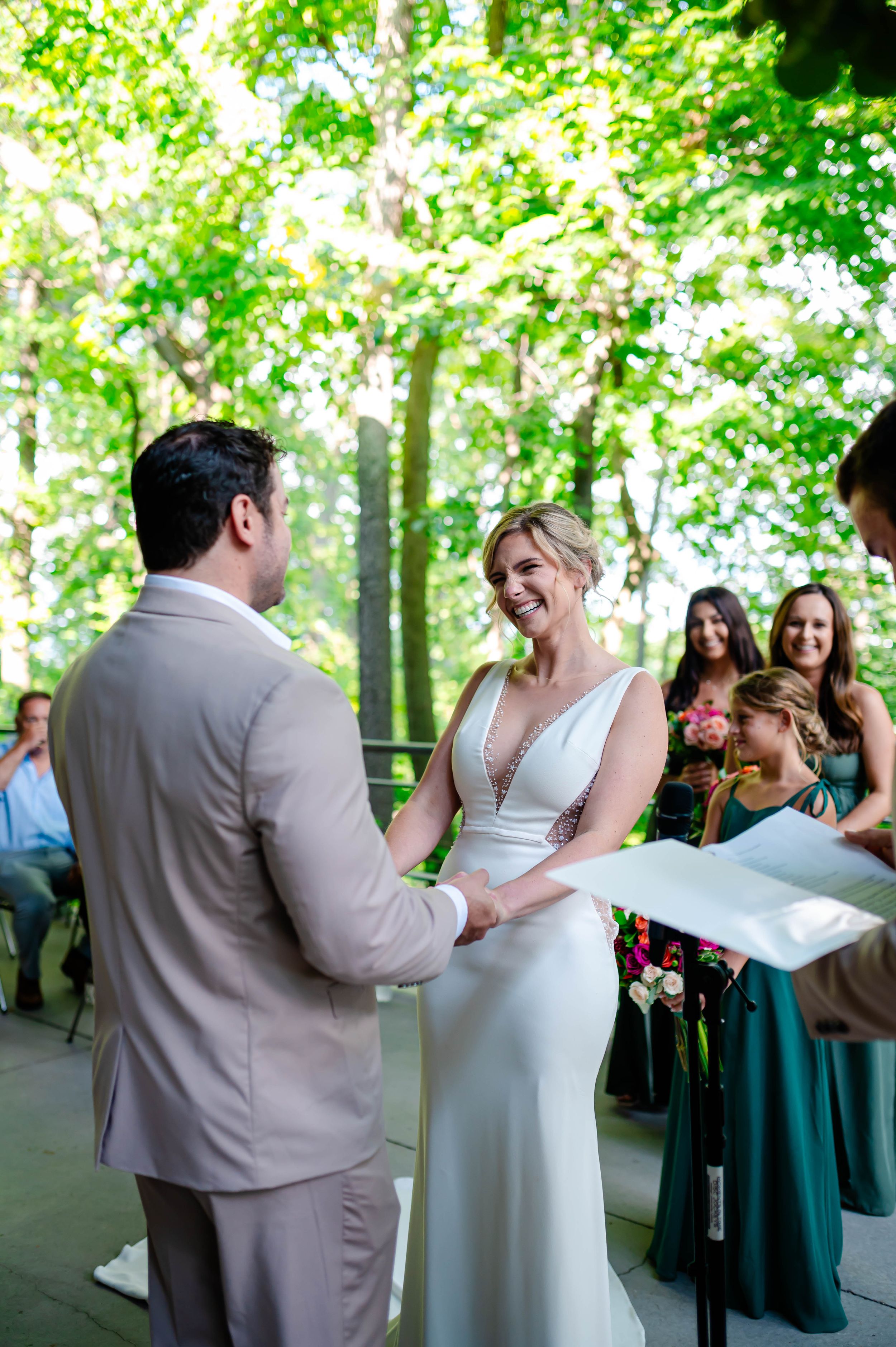 Bride laughs joyfully holding hands with groom during outdoor wedding ceremony surrounded by guests.