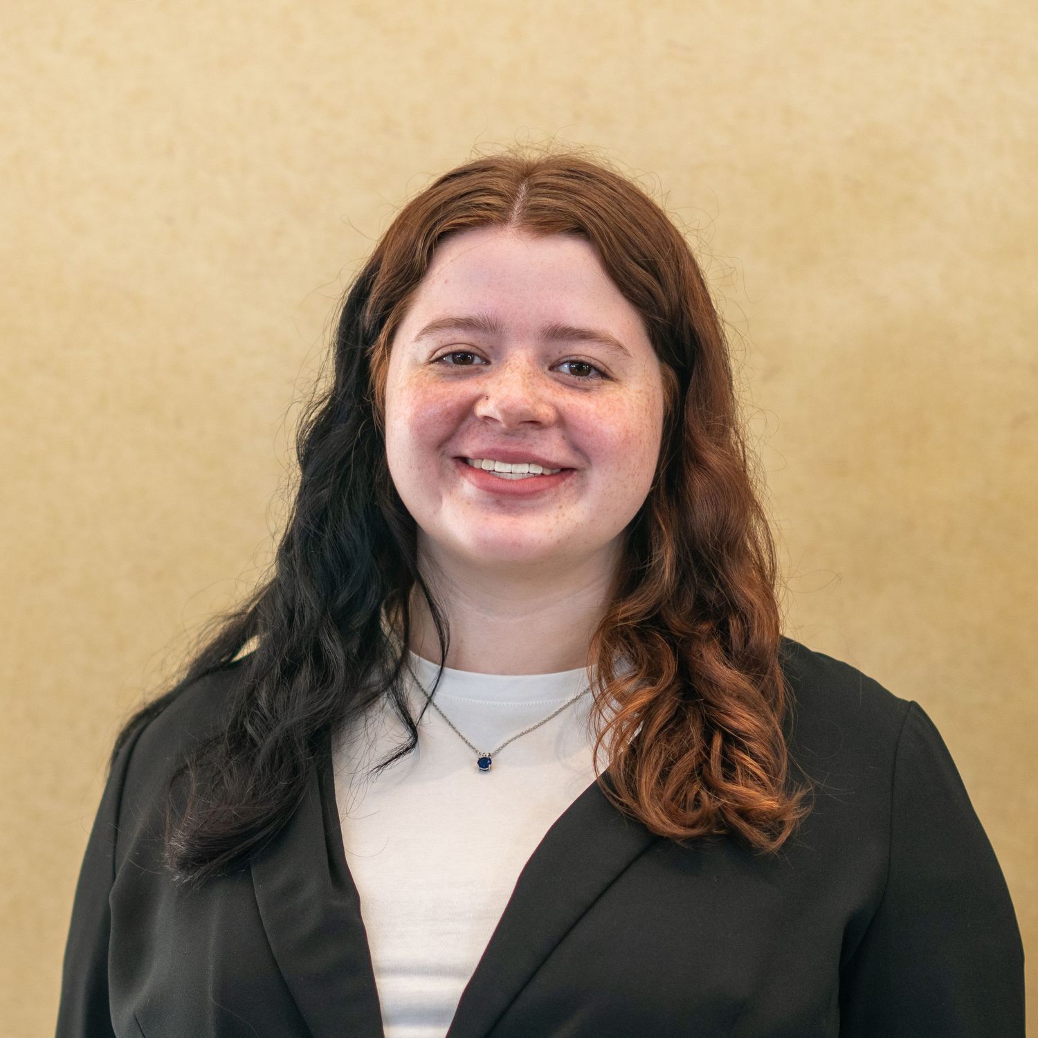 Young professional woman with red hair smiling in a black blazer against a neutral background.