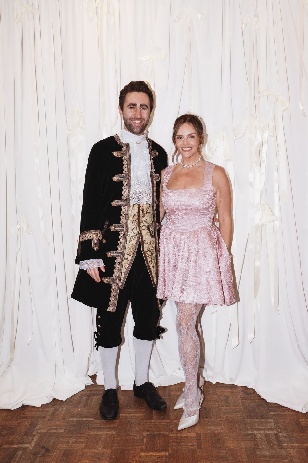 Couple posing in elegant period costumes, man in black baroque jacket, woman in pink mini dress with lace tights.