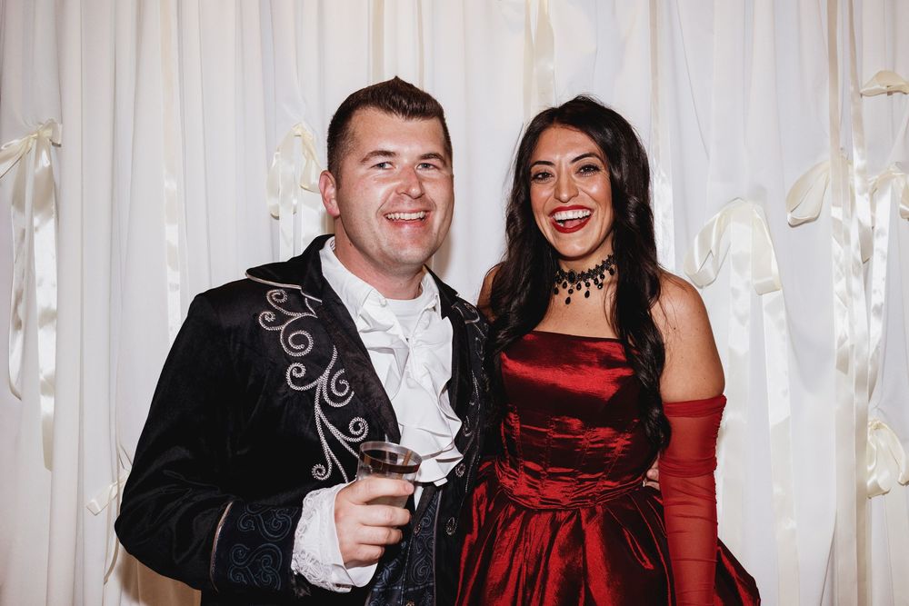 Elegant couple in formal attire posing together, man in black embroidered suit, woman in red satin gown with gloves.