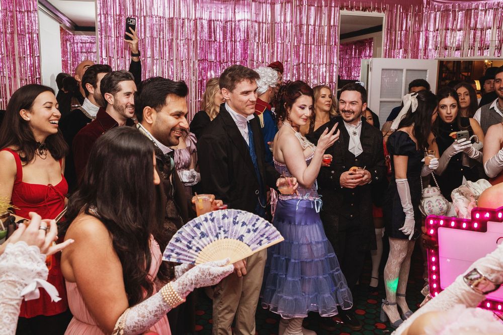Guests in formal attire dance and socialize at a festive party with pink tinsel backdrop and elegant decorations.