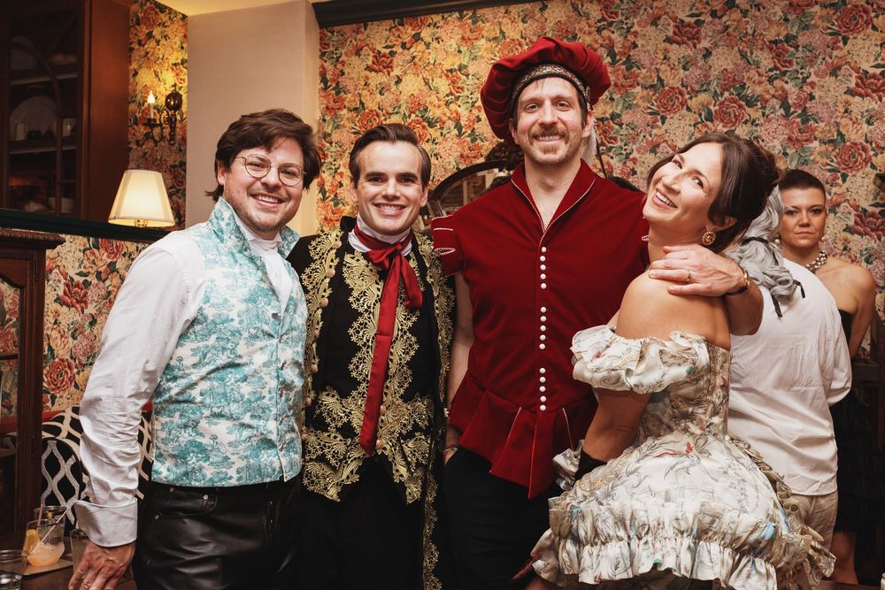 Four performers in elegant Victorian costumes smile together at a formal themed event or theatrical production.
