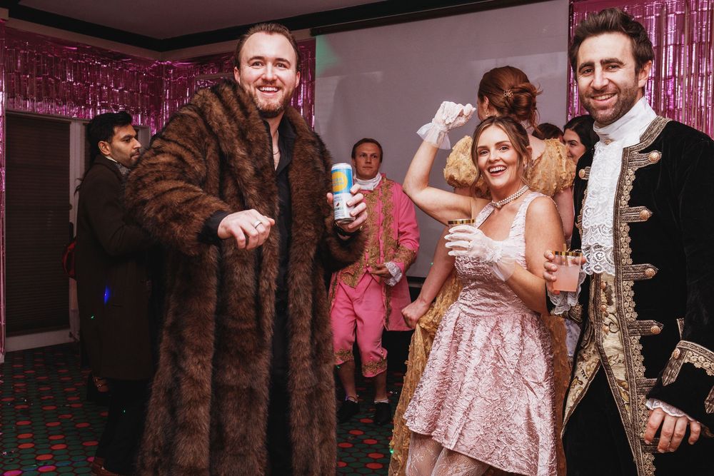 Three adults in fancy dress costumes dancing and posing at a glamorous themed party with pink decorations.