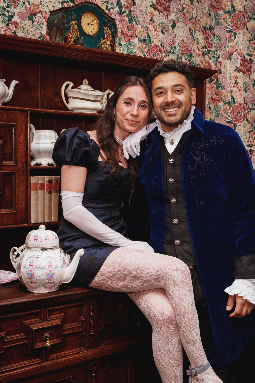 Couple in vintage Victorian costumes posing by antique dresser, woman in navy dress with white gloves, man in blue velvet coat.