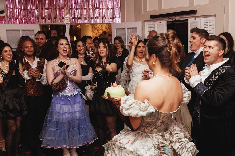 Guests celebrate with birthday cake at elegant party, woman in strapless gown holds cake while crowd cheers.