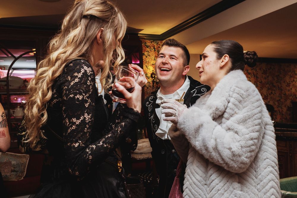 Three guests laughing and socializing at upscale party, woman in lace dress and man in embroidered jacket.