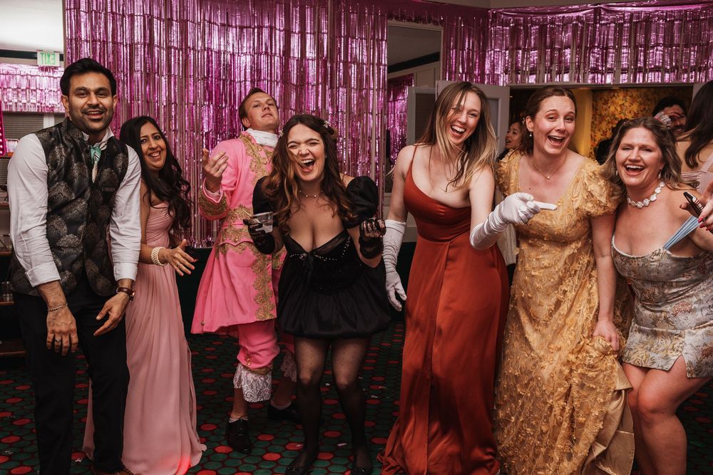 Group of friends dancing and smiling at a glamorous party with pink tinsel backdrop and formal dresses.