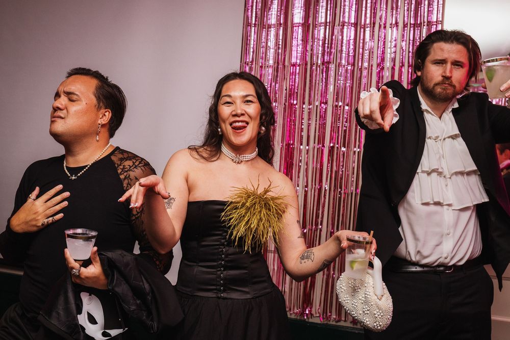 Three friends pose playfully at a festive party with pink tinsel backdrop, wearing costumes including a vampire outfit.