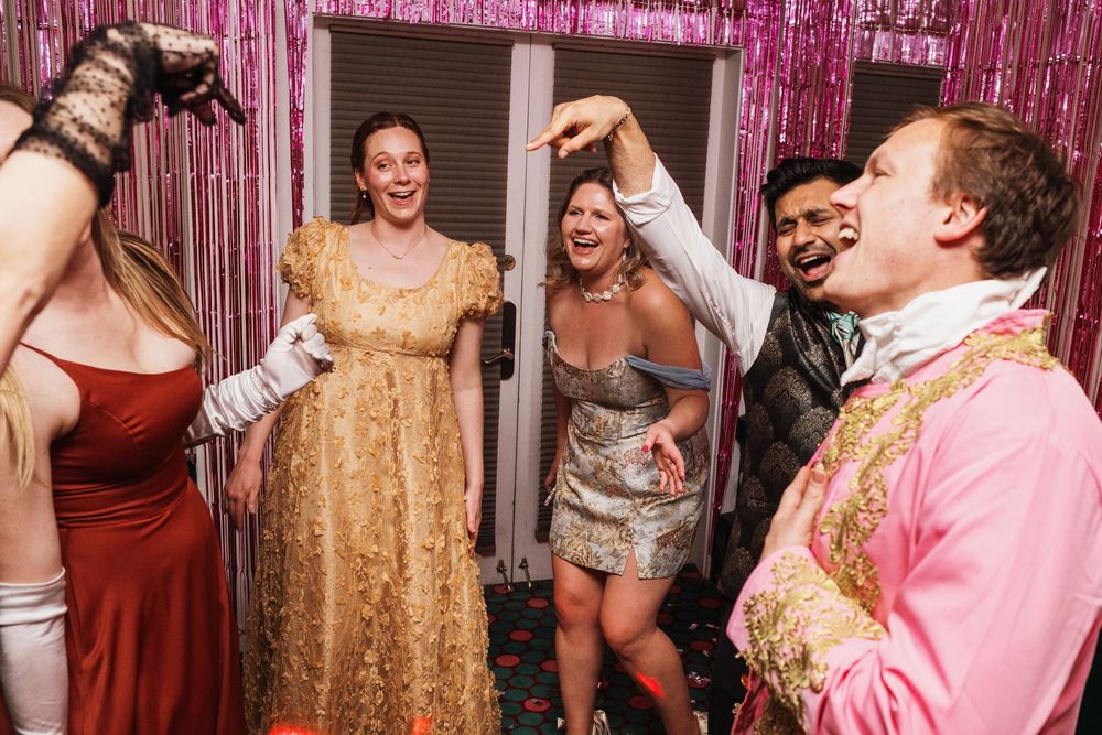 Group of friends laughing and dancing together at a glamorous costume party with pink tinsel backdrop.