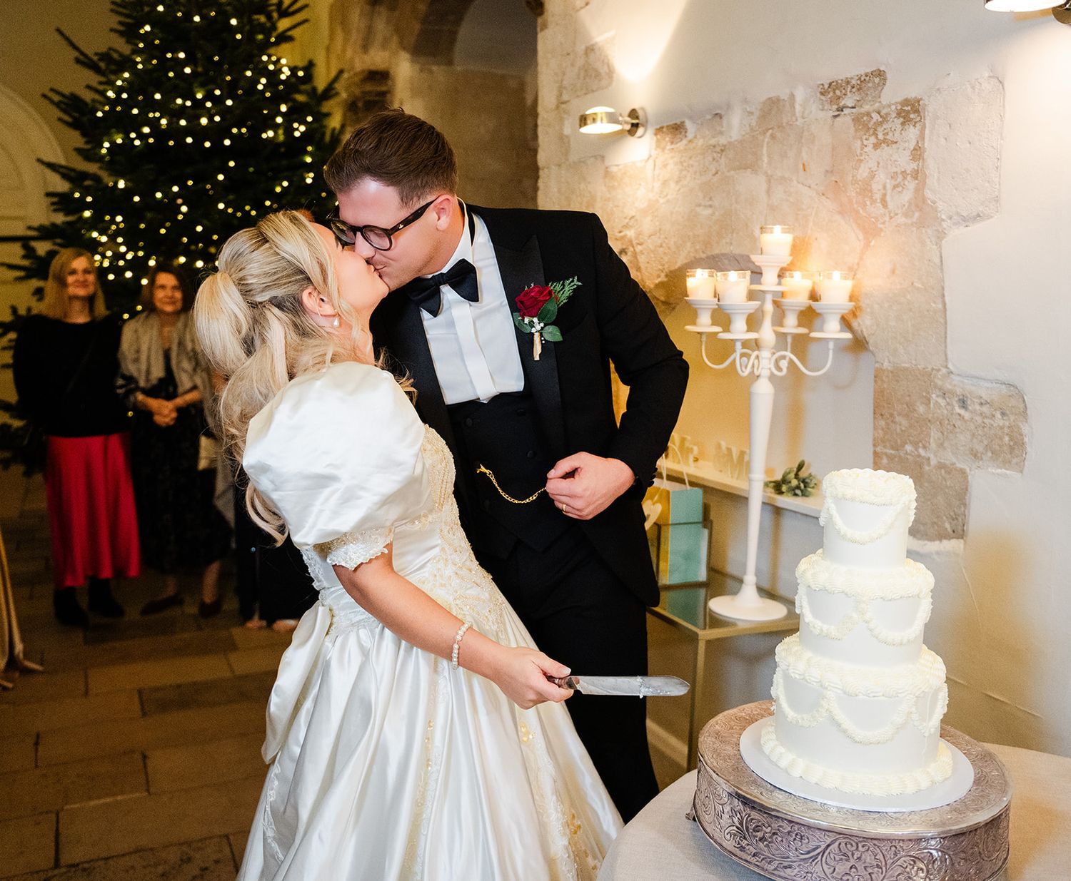Bride and groom sharing a kiss while cutting their white tiered wedding cake at a Christmas-themed reception.