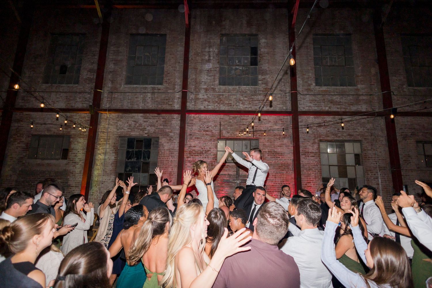 Lively wedding reception dance floor crowd in rustic brick venue with string lights and guests raising hands.