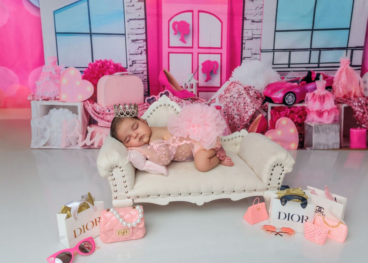 Baby girl in pink tutu sleeps on white sofa surrounded by Dior accessories in Barbie-themed setup.