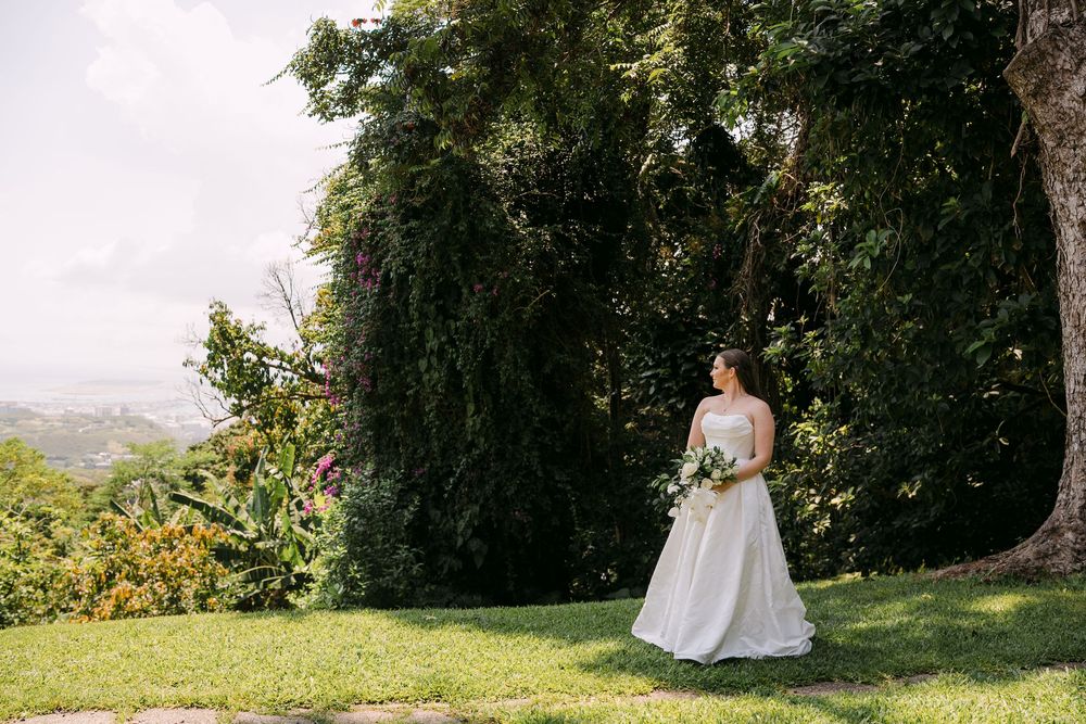 Bride in white gown holding bouquet stands on lush green lawn surrounded by tropical trees with ocean view.